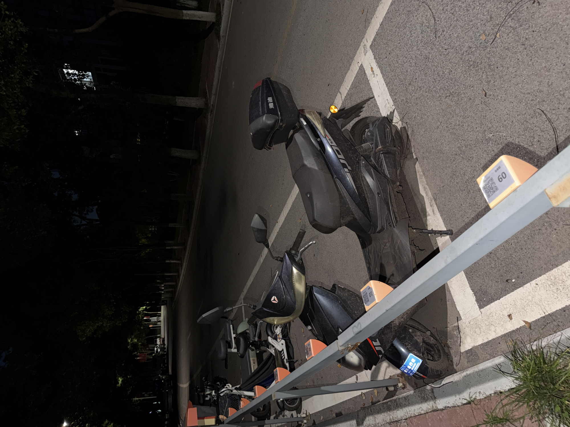 In the city of Jiangjin, China, at night, a row of parked electric scooters is visible along a street. A gray metal railing separates the scooters from the sidewalk. The scooters are parked in designated spots marked by white lines on the asphalt. One scooter in the foreground is black with a black storage box on the back. It has an orange tag with a QR code and the number "60" on its front. Another scooter to its left is white and blue, also with an orange tag and a QR code. In the background, the street is lined with trees that cast dark shadows. The streetlights illuminate the road and the area where the scooters are parked. A few cars are parked on the opposite side of the street. The overall lighting suggests it is nighttime with artificial light sources.