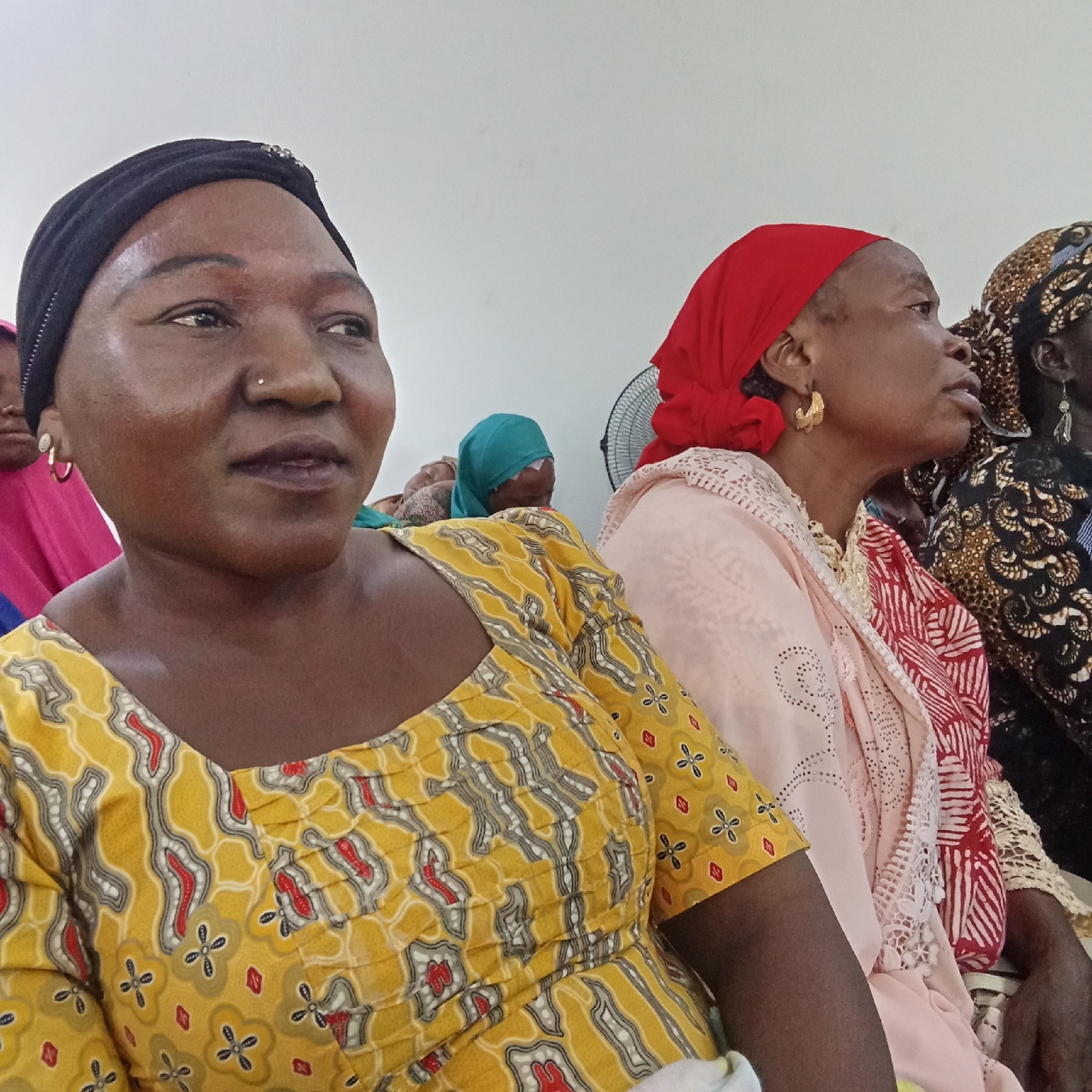 The scene depicts multiple women seated indoors in Akwanga, Nigeria. In the foreground, a woman with dark skin wears a black headwrap adorned with silver accents, a small silver nose stud, and gold-colored hoop earrings. She is dressed in a yellow garment featuring a repeating pattern of grey, red, and white abstract shapes and floral motifs. Her gaze is directed slightly upward and to the right. Behind her and to the right, a second woman with dark skin wears a red headwrap and large, textured gold-colored hoop earrings. She has a light pink lace-trimmed garment over what appears to be a red and white patterned fabric. Her head is turned to her right. Partially visible further in the background are additional women, one with a teal headwrap and another with a dark brown and gold patterned headwrap and garment. A black oscillating fan is visible to the right of the second woman's head. The background consists of a plain white wall. The women appear to be seated in rows, indicative of a gathering or meeting.