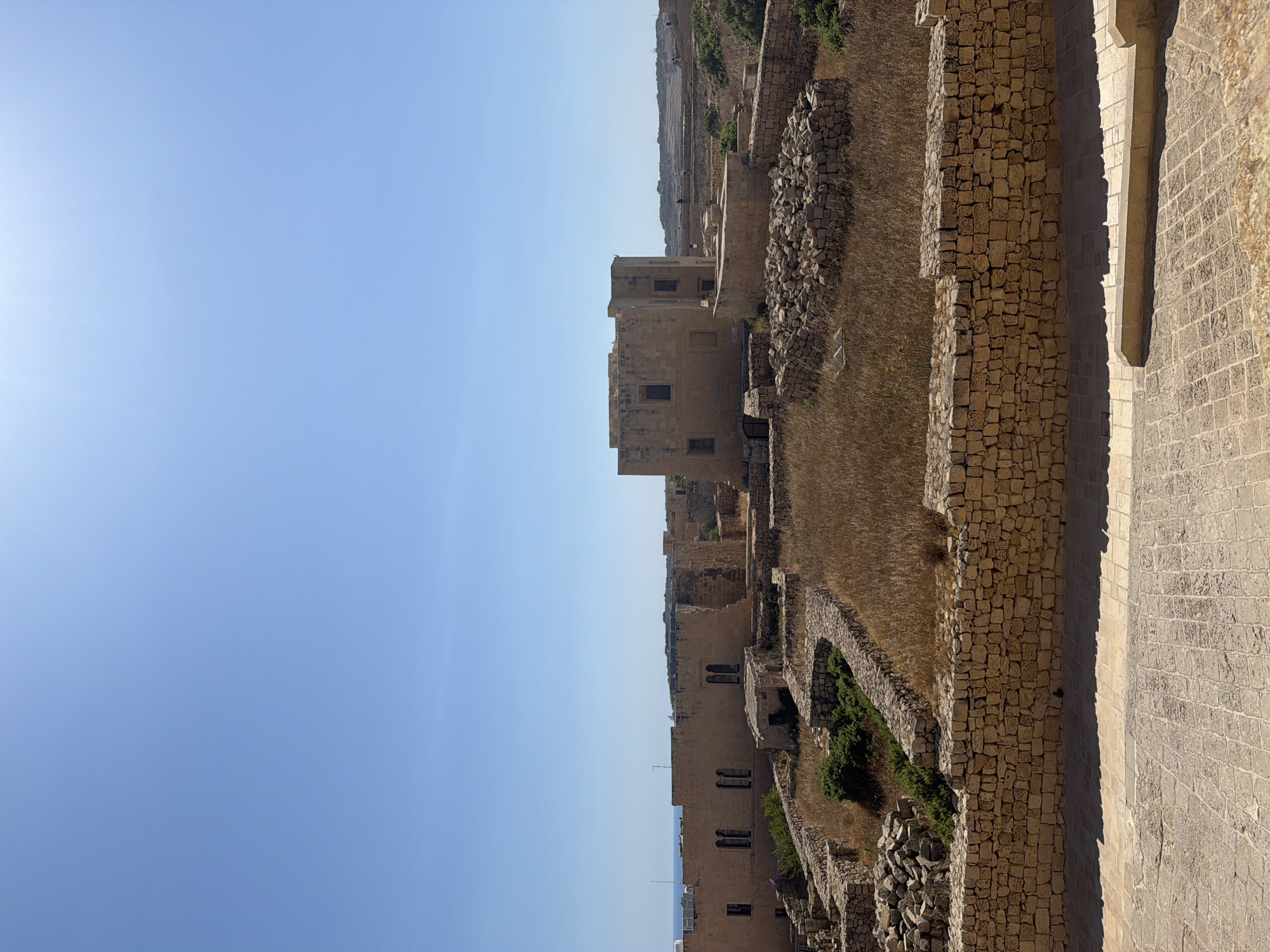 The image displays the exterior of the Citadel in Victoria, Gozo, Malta. The scene is bathed in bright sunlight under a clear blue sky. In the foreground, a paved walkway made of light-colored cobblestones runs horizontally, casting distinct shadows. A low, stone wall constructed from irregular, light brown stones separates the walkway from the interior of the Citadel complex.
Beyond this wall, the ground is covered in dry, golden grass and scattered stones, suggesting a somewhat arid environment. Various stone structures, appearing to be ruins or historical buildings, are visible. These include low walls outlining different sections and a more substantial, light brown stone building with visible windows, possibly a tower or a central structure within the complex. Another, slightly taller stone building with multiple windows is also prominent, with a flat roof and visible ventilation shafts. In the distance, more ruins and the faint outline of land and sea can be seen. There are a few figures walking in the background, too small to discern details. The overall impression is one of a historic, sun-drenched archaeological site.