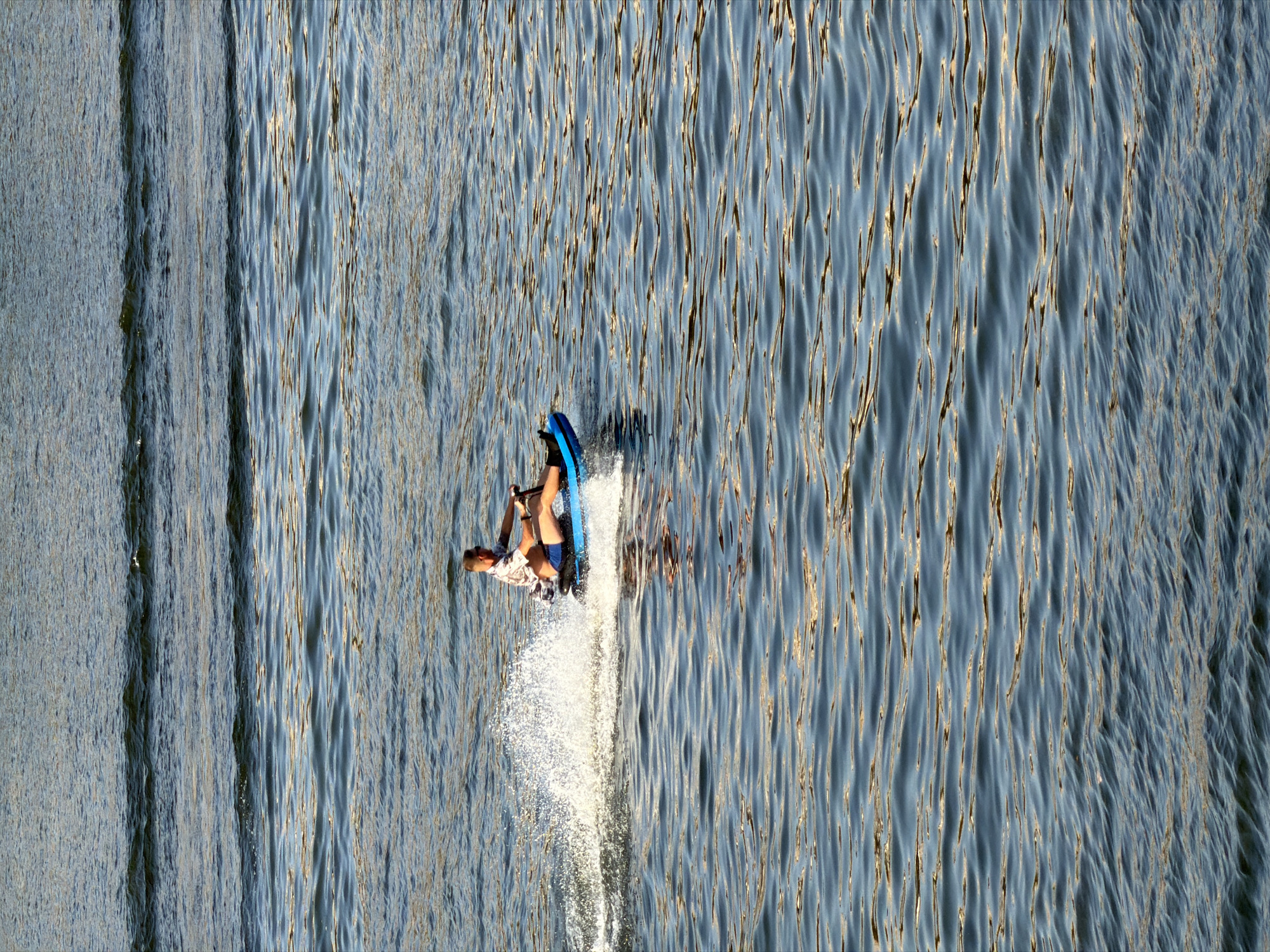 A person rides a blue watercraft on a body of water, leaving a wake. The person is wearing a patterned short-sleeved shirt and blue shorts and is holding onto handlebars. The watercraft has a black seat. The scene appears to be a sunny day, with sunlight reflecting off the rippling water. This image was taken on the Volga River near Kamyshin, Russia.