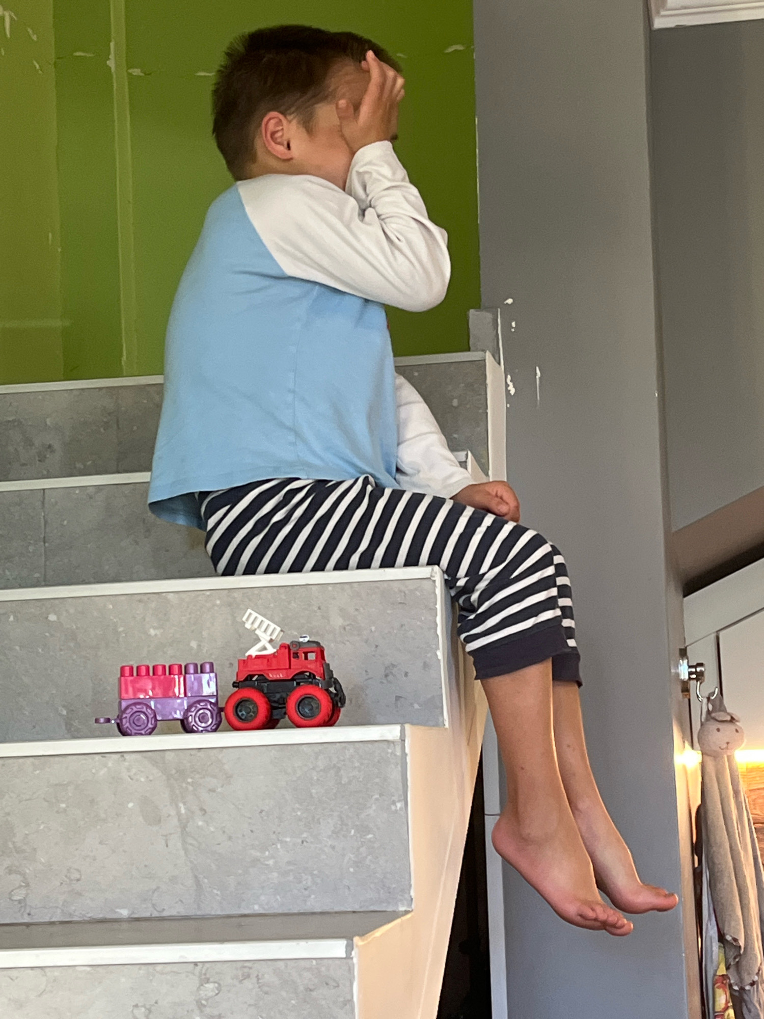 A young boy is sitting on a concrete staircase, his legs dangling off the side. He is wearing a long-sleeved shirt with a light blue top and white sleeves, and striped pants with navy blue and white stripes. The boy has his hand over his face, as if he is hiding or feeling embarrassed. On the step below him are two toy vehicles: a purple train car and a red fire truck. The background features a bright green wall and a gray wall. The scene appears to be indoors, possibly in a residential setting. The image does not provide any specific information to place the location in Ureki, Georgia.