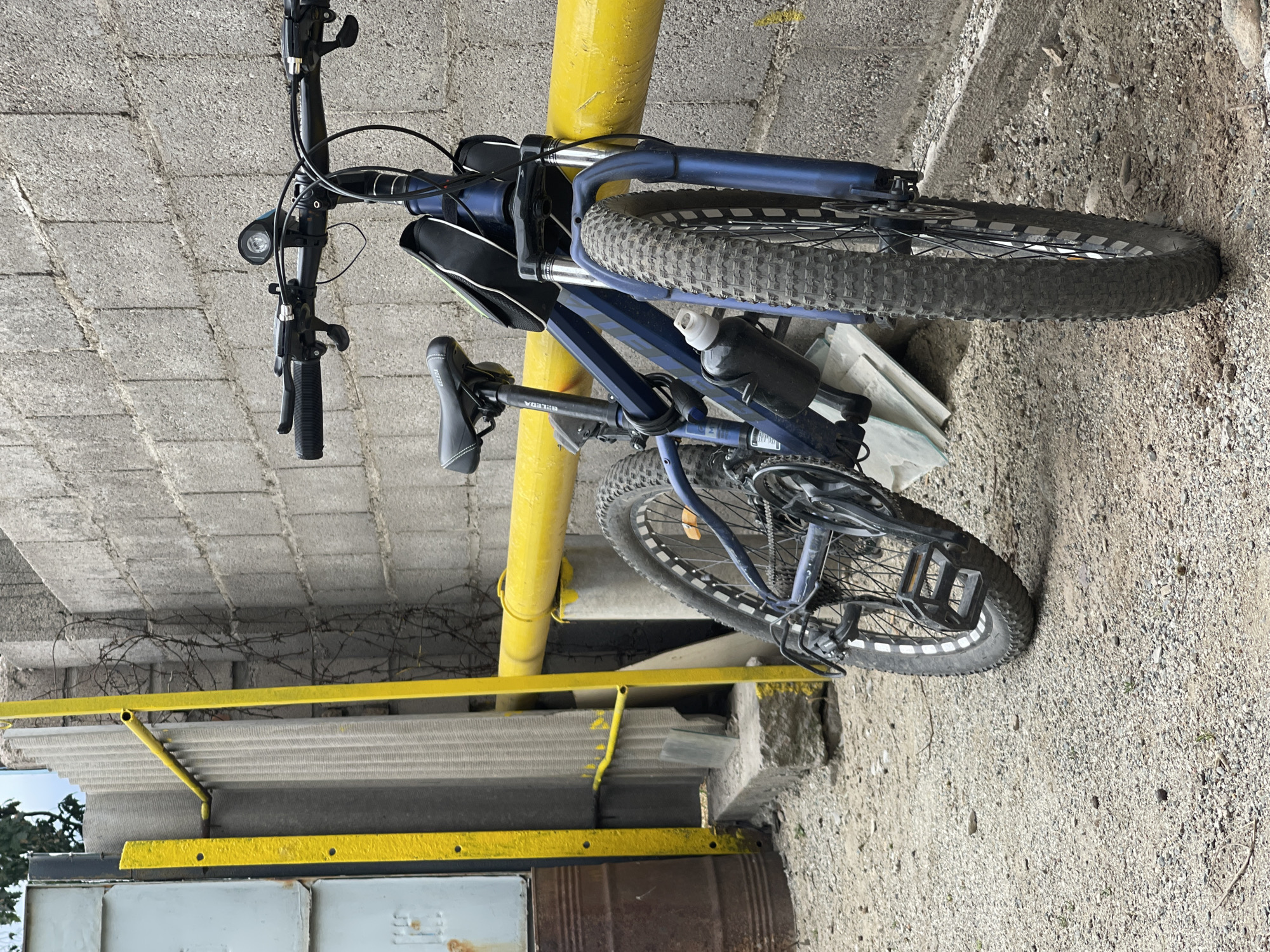 The image shows a blue fat-tire bicycle leaning against a yellow pipe. The bicycle has thick tires with white accents, a black seat, and a black water bottle cage attached to the frame. A black saddle bag is also attached to the bicycle. The bicycle is parked in an outdoor area with a concrete block wall in the background and a gravel-covered ground. There is barbed wire visible above the bicycle, and a corrugated metal roof structure is present to the left.
The scene appears to be in Bishkek City, Kyrgyzstan, suggested by the mention of the location context. The lighting suggests it is daytime. The environment seems to be somewhat industrial or utilitarian, with exposed pipes and concrete structures. No people are visible in the image. The bicycle is the primary subject, and it appears to be stationary. No specific activity is taking place, beyond the bicycle being parked. There is no visible text on the bicycle or in the surrounding environment.