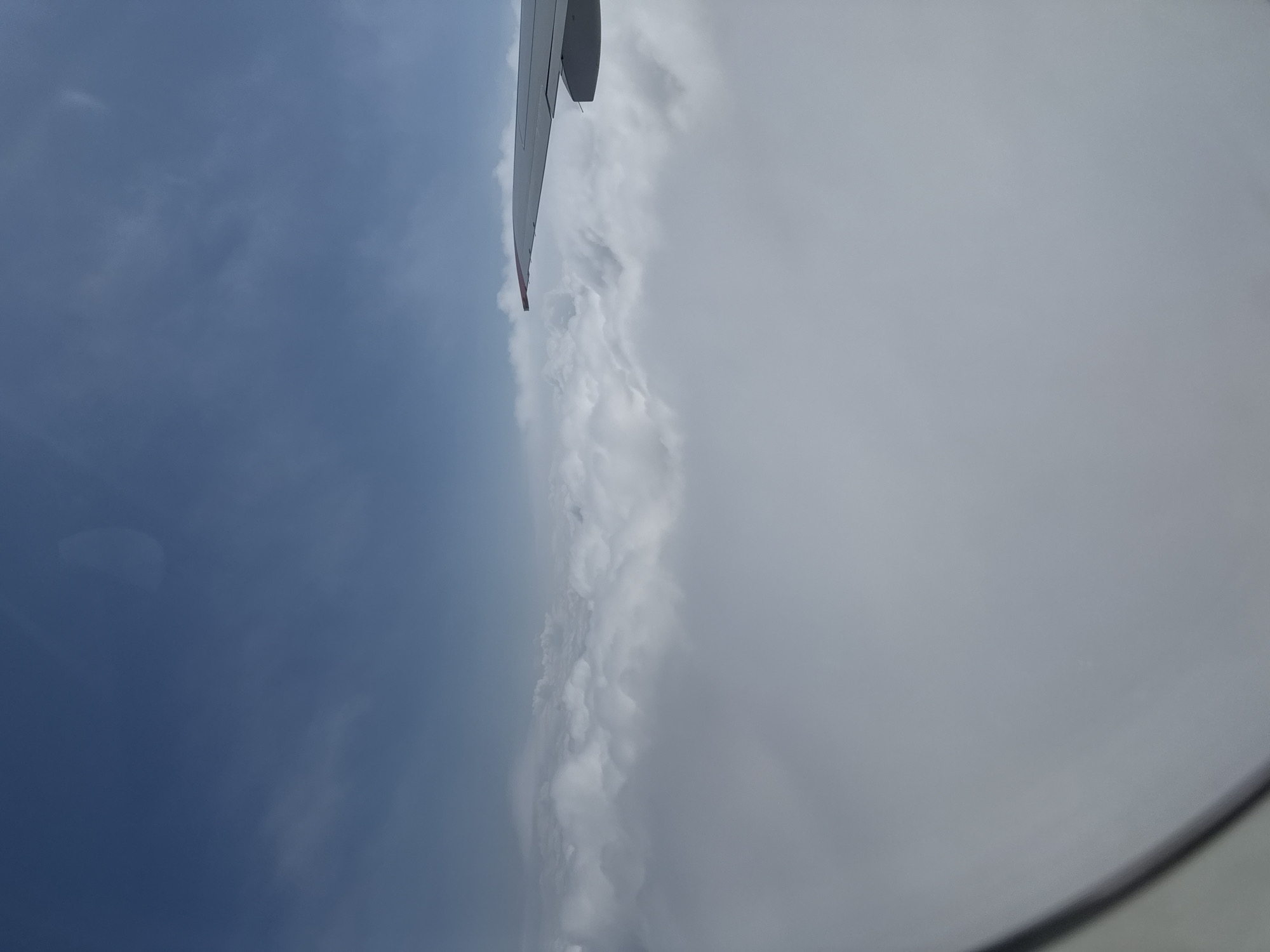 An aerial photograph taken from an airplane window shows a view of clouds and a portion of an airplane wing against a blue sky. The clouds are dense and white, forming a thick layer below the aircraft. The wing is visible in the upper right corner of the frame, angled towards the clouds. The sky is a clear blue with some wispy clouds scattered throughout. The image does not provide any information about the city of Staroe Mart'yanovo, Russia, or any people.