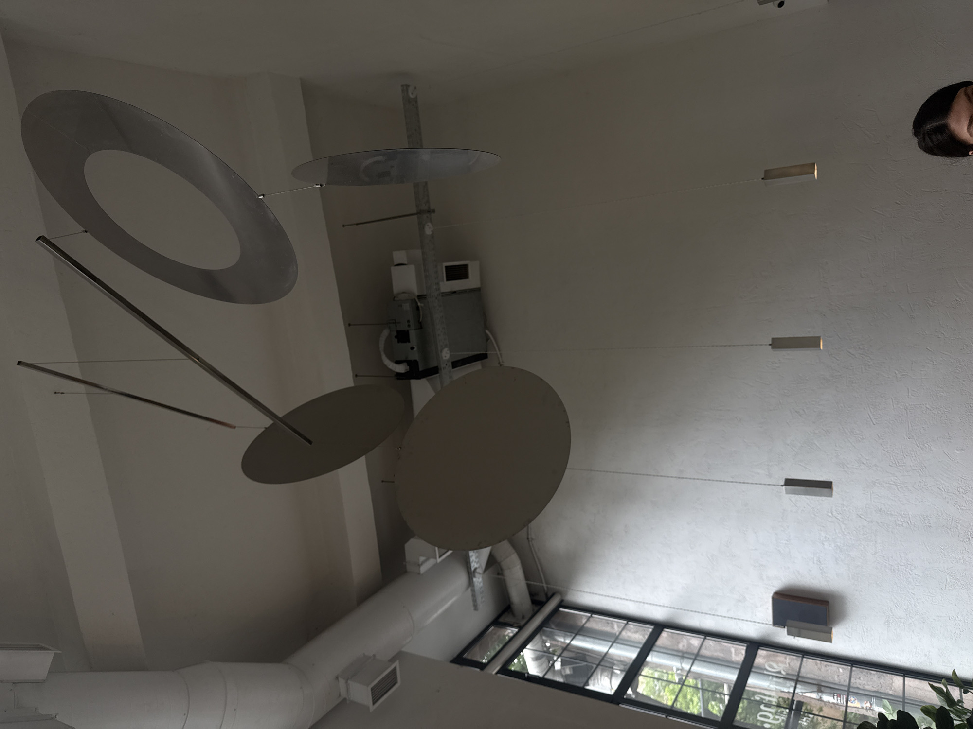 The image depicts an interior ceiling with a complex art installation of metal shapes, including a circular ring, smaller circles, and supporting rods. The ceiling is white and features recessed lighting fixtures connected by thin wires. An industrial-style air duct is also visible. Part of a face can be seen in the upper right corner of the image. The lower part of the image shows windows offering a glimpse of greenery. The image appears to be taken indoors in Yerevan, Armenia.