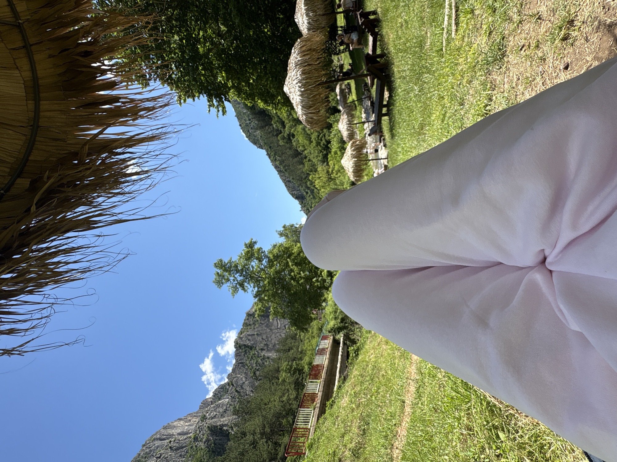 The image captures a serene outdoor scene in Zgorigrad, Bulgaria, likely from a seated perspective under a thatched umbrella. In the foreground, the lower half of a person's legs, clad in light pink pants, dominate the view. The background unfolds into a lush green landscape with a river meandering through it. Several straw umbrellas and lounge chairs are set up on the grass, suggesting a recreational area. Beyond the grassy area, a gently sloping hillside covered in trees rises, leading up to imposing, rugged mountains that form the backdrop. A small bridge with red and white railings is visible on the left side of the frame, spanning across a grassy incline. The sky is a clear, bright blue with a few wispy white clouds. The overall impression is one of tranquility and relaxation in a natural setting.