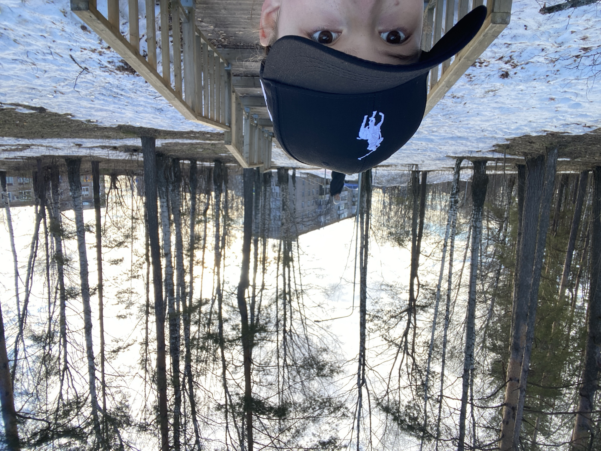 The image shows a person wearing a black baseball cap with a white polo player logo, viewed from an upside-down perspective. The background reveals a snowy landscape with trees and a building, likely taken from a bridge or elevated structure. The location is inferred to be Chaikovsky, Russia based on the buildings and environment. Details include snow-covered ground with bare trees, a wooden railing on the bridge, and a multi-story building in the distance. The subject is looking directly at the camera.