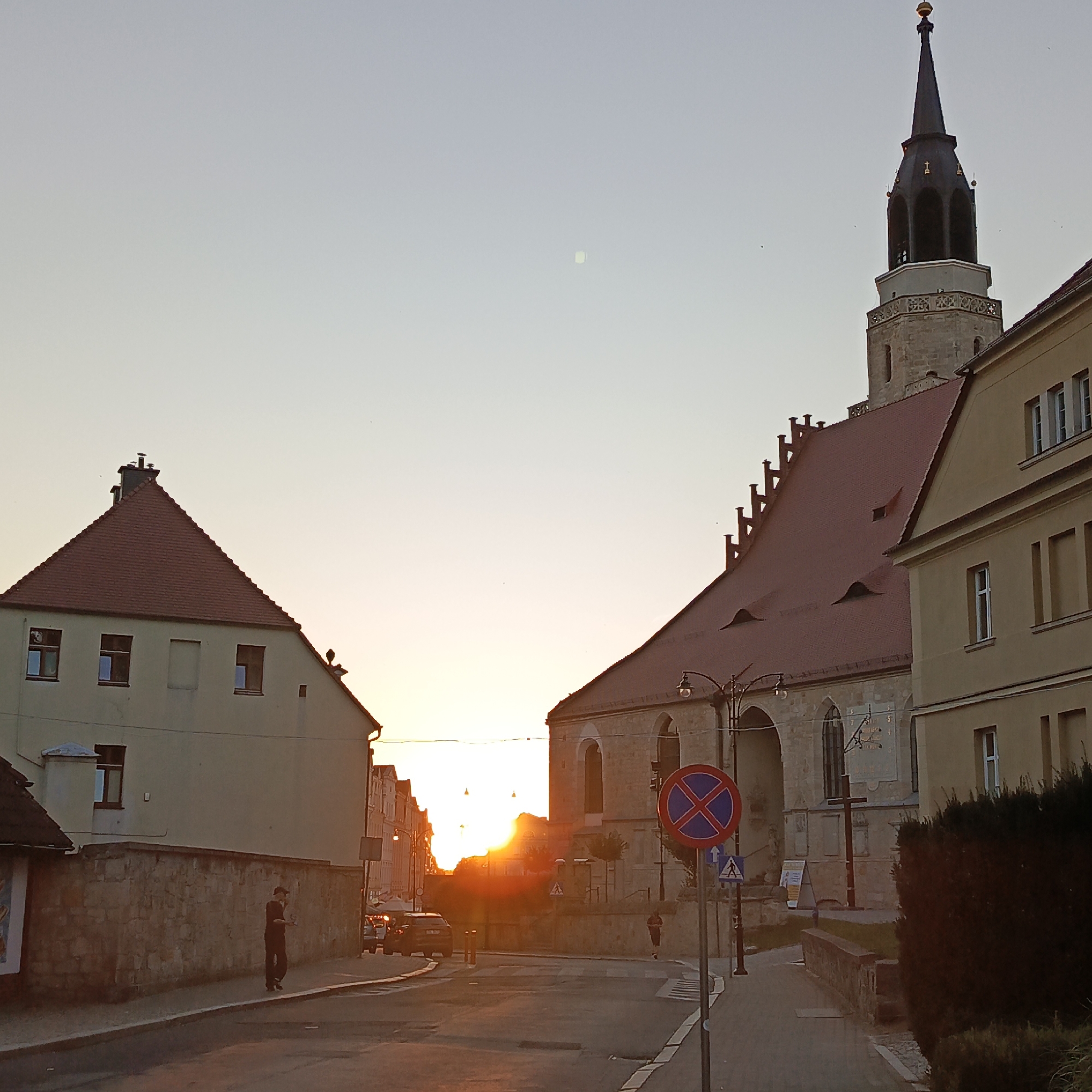 The image depicts a street scene in Bolesławiec, Poland, at sunset. The sun is low on the horizon, casting a warm glow over the scene and creating lens flare. On the right side of the image, a prominent church tower with a dark spire and a golden finial stands tall against the pale sky. The church itself has a light-colored stone facade with arched windows and a red tiled roof. Adjacent to the church, a yellow building with multiple windows and a red tiled roof is visible.
On the left side of the street, a lighter-colored building with a red tiled roof and several windows is present. A stone wall runs along the sidewalk in front of this building. A person dressed in dark clothing is walking along the sidewalk near this wall.
The street itself is paved and appears to be a main thoroughfare in the town. Several cars are visible on the street, including a dark-colored SUV. A "no parking" sign with a red circle and blue cross is prominently displayed on a pole near the church. Another pedestrian crossing sign is also visible. A street lamp with decorative metalwork can be seen to the left of the church. The overall atmosphere of the image is tranquil, characteristic of a European town at dusk.