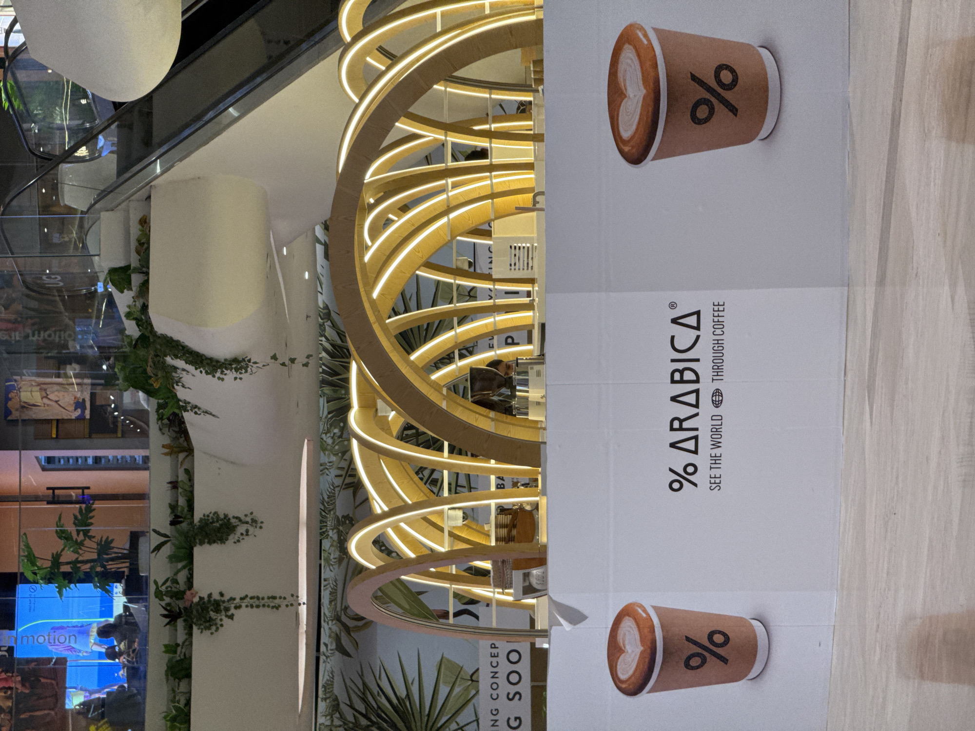 A modern, stylish coffee shop named "% ARABICA" is featured prominently within what appears to be an upscale shopping mall in Denpasar, Indonesia. The coffee shop boasts a distinctive architectural design characterized by multiple glowing, curved wooden or light-colored bands that form a spiral or wave-like structure, creating an eye-catching facade. Lush green plants and vines are strategically placed around the base and pillars of the shop, adding a natural touch to the contemporary design.
A large white vertical sign on the right side of the image displays the coffee shop's brand name, "% ARABICA," along with its tagline, "SEE THE WORLD THROUGH COFFEE." The sign also features two illustrations of coffee cups, each adorned with latte art in the shape of a heart and the brand's unique "%" logo.
In the background, an escalator is visible, reflecting the bright indoor lighting. Other elements suggesting a commercial interior include polished floors and a reflective surface, possibly glass, showcasing parts of the mall environment. On a screen further in the background, a blue light emanates with the word "motion" partially visible. Another partial sign on the bottom left reads "NG CONCEP G SOO," likely indicating "COMING SOON" or "NEW CONCEPT," suggesting a recent or upcoming opening for this specific outlet. The scene is brightly lit, indicative of daytime or a well-lit indoor environment, exuding a clean, inviting, and modern atmosphere.