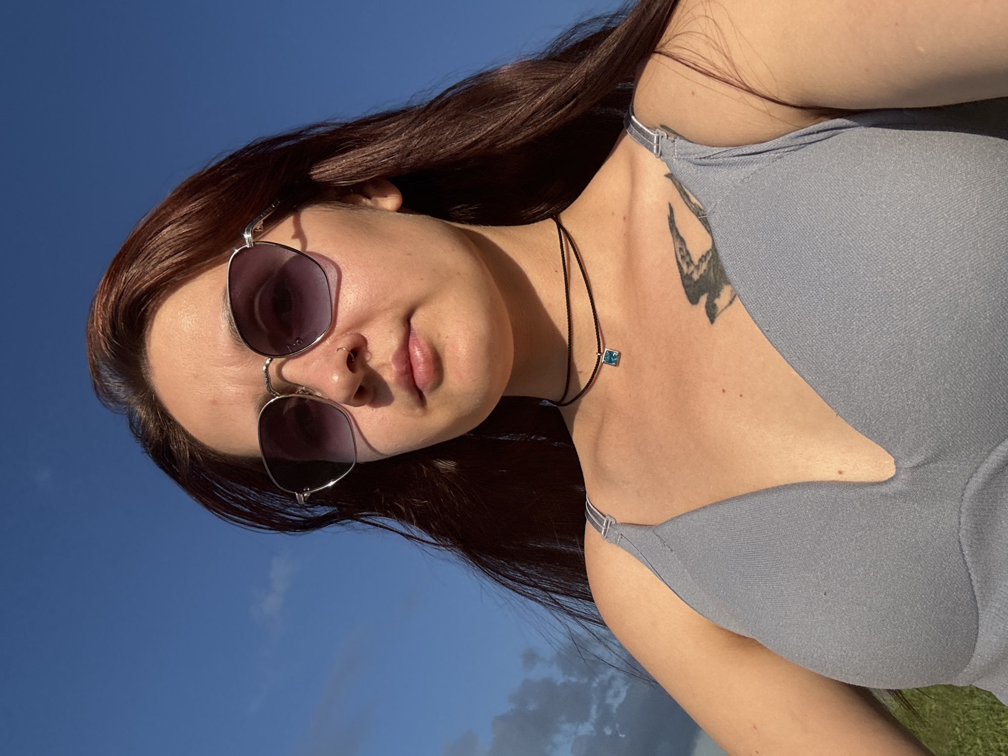 In the city of Naberezhnye Chelny, Russia, a young woman with long, dark brown hair is captured in a low-angle selfie. She is wearing a grey top with thin straps and a necklace with a blue pendant. Her face is partially obscured by sunglasses with purple lenses. A tattoo of a bird is visible on her upper chest. The background consists of a bright blue sky with a few scattered clouds. The lighting suggests it is daytime, possibly late afternoon due to the shadows cast by the sunglasses. The woman's expression is neutral as she looks towards the camera.
