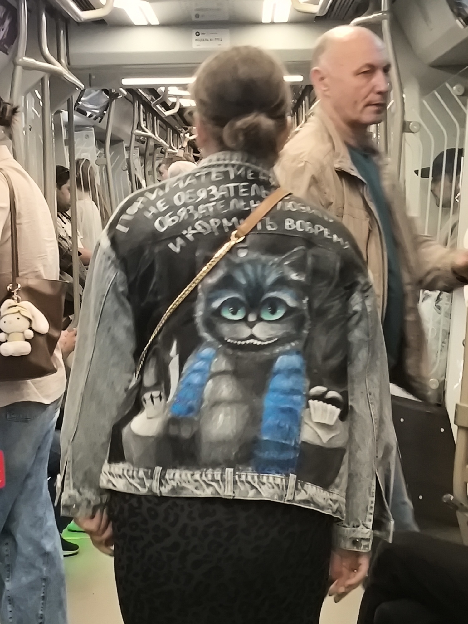 The image shows the back of a person wearing a denim jacket with a painted design of a cat with green eyes. White text is visible on the jacket above the cat image. The person is inside a public transport vehicle, likely a train, with other passengers visible in the background. The environment appears to be urban, with metal poles and seats characteristic of public transportation. The lighting is artificial and somewhat dim.