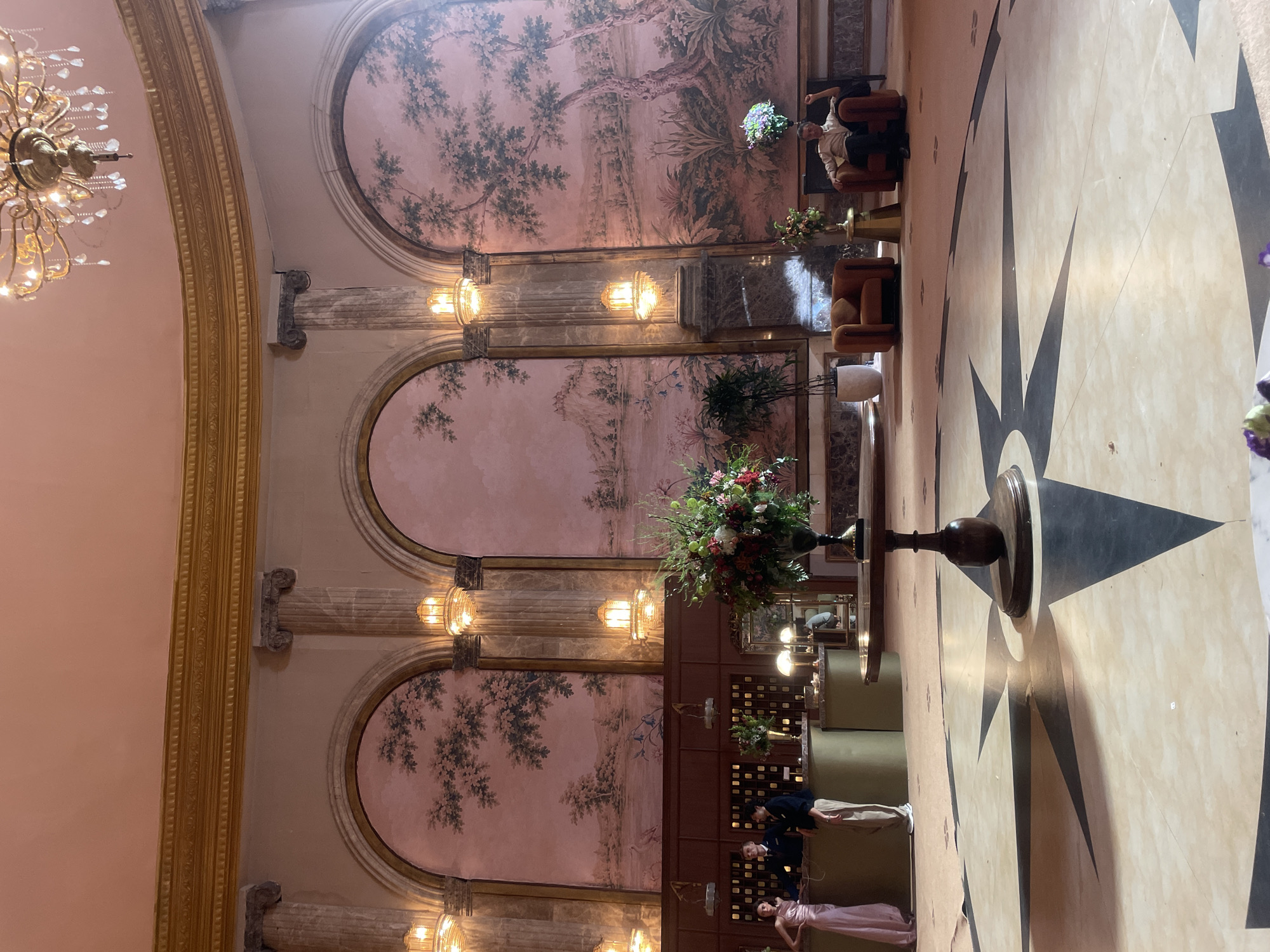 The image depicts the grand lobby of the Taj Mahal Palace Hotel in Mumbai, India. The spacious lobby features a high, ornate ceiling with a large chandelier. The walls are adorned with three large arched murals depicting pastoral landscapes. Two classical columns flank the central mural, and smaller arched alcoves with decorative lighting are visible on either side of the main arches.
In the foreground, a large, round table with a wooden base holds a substantial floral arrangement of red, green, and white flowers. The floor is a patterned marble inlay, with a prominent starburst design in the center.
To the left, a reception desk is staffed by two individuals. A woman in a pale pink gown stands to the left, and two men in business attire stand behind the counter. A woman can be seen through a mirror behind the counter.
To the right, two plush, burnt-orange armchairs are placed near the wall. A man in a light-colored shirt and dark pants is seated in the right armchair, with his right arm raised. A small side table with a gold finish is positioned between the armchairs, holding a small floral arrangement.
The overall impression is one of opulent grandeur and classic elegance, characteristic of a historic luxury hotel.