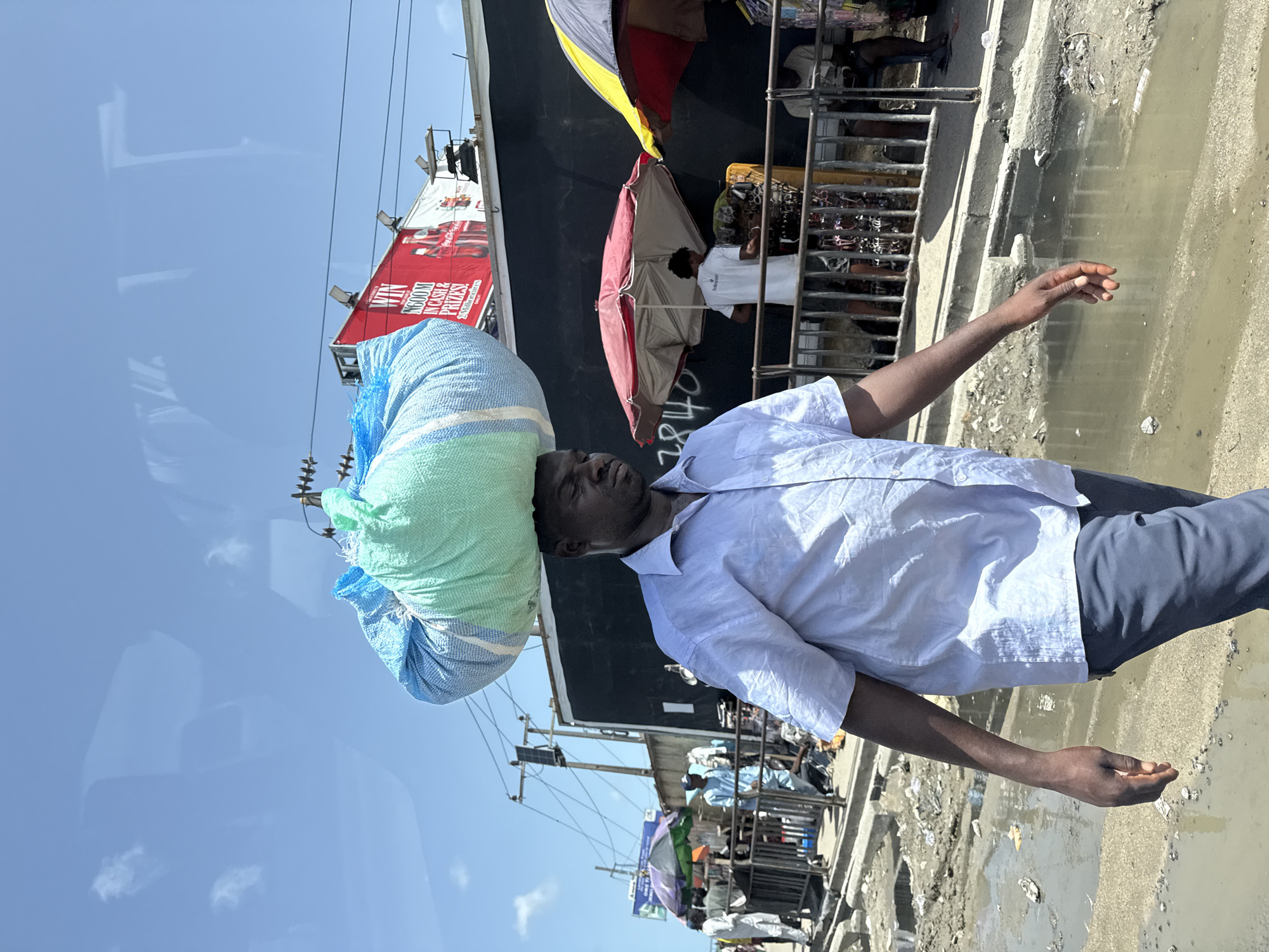 A man is carrying a large blue and green sack on his head. He is wearing a white shirt and dark pants. The background shows a busy Lagos street scene, Nigeria. There is a Coca-Cola advertisement, various stalls, umbrellas, and people. The ground is wet and looks like a street market.