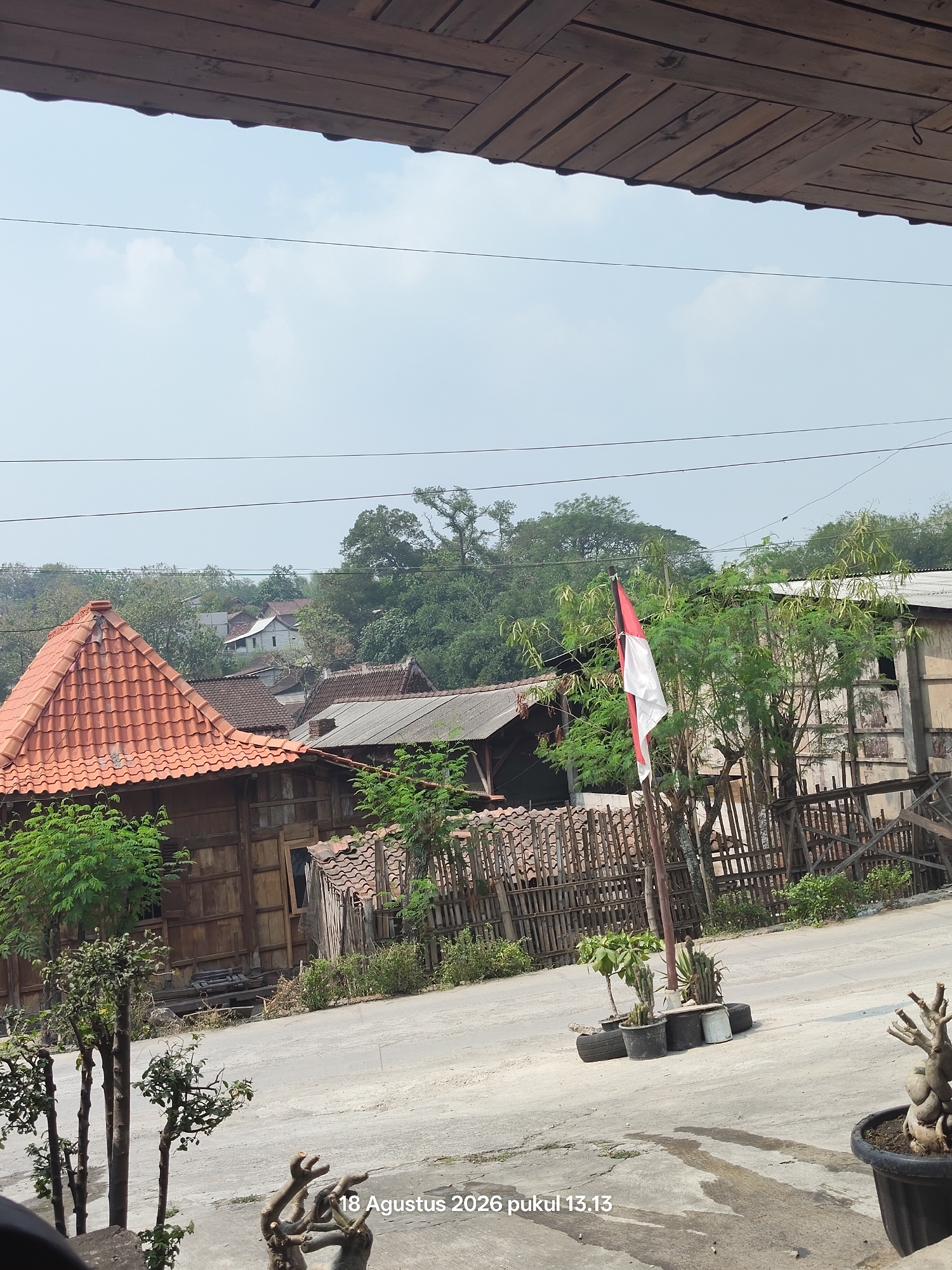 This is a daytime, eye-level, medium shot of a village scene in Sugihmanik, Indonesia. The foreground is dominated by a paved road with several potted plants, including cacti, arranged on the side. To the left, bare tree branches are visible, and to the right, a potted plant with a gnarled trunk sits in a black pot. Further back, a wooden fence encloses an area with more greenery and some rudimentary structures. A red and white Indonesian flag is prominently displayed on a pole in front of a building with a weathered wooden facade and a light-colored roof. Several other buildings with tiled roofs and wooden walls are scattered throughout the background, nestled amongst lush green trees and foliage. The sky is a pale blue with scattered clouds. Text overlayed on the bottom of the image reads "18 Agustus 2026 pukul 13.13".