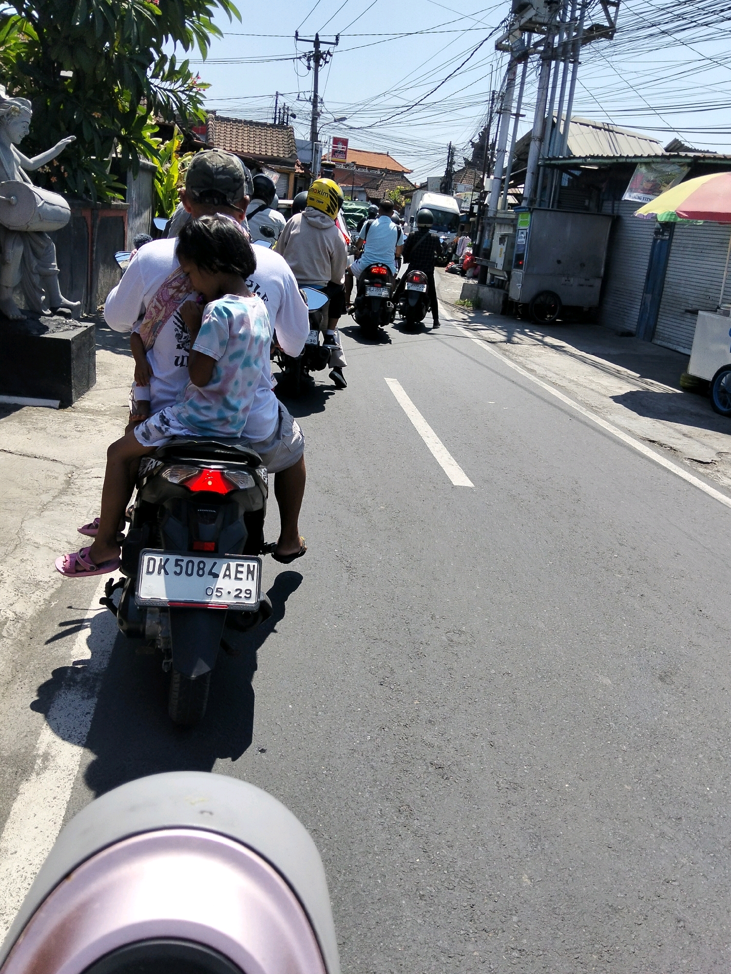 In Denpasar, Indonesia, a man wearing a camouflage baseball cap and a white t-shirt rides a black motorcycle. He is carrying a young child, who is wearing a white and pink tie-dye t-shirt. The child has their arms wrapped around the man's neck. Both are wearing casual clothing suitable for a warm climate. The motorcycle has a license plate that reads "DK 5084 AEN". The scene takes place on a paved road with a white line marking the lane. In the background, there are buildings, utility poles with numerous wires, and other motorcycles and a white truck. A statue of a musician can be seen on the left side of the image. A red and yellow umbrella is visible on the right. The overall environment suggests a busy street in a tropical urban setting.