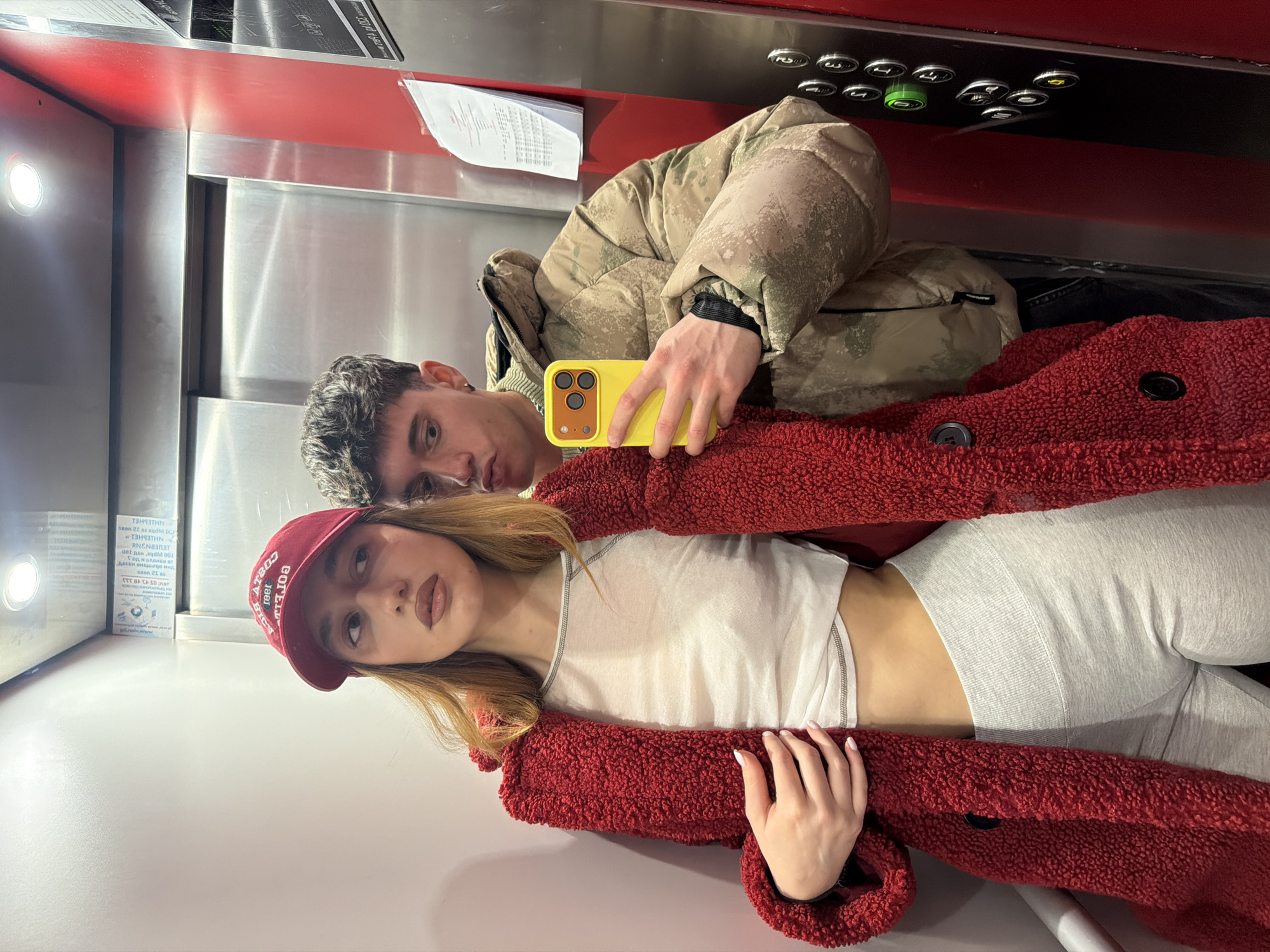 A young man and woman are in an elevator. The man, dressed in a beige camouflage puffer jacket, holds a yellow iPhone and appears to be taking a selfie with the woman, who is positioned in front of him. The woman is wearing a red "COSTA" baseball cap, a white crop top, grey leggings, and a large red faux fur jacket. The elevator interior features stainless steel walls, a red panel at the top with elevator buttons, and a light. A printed document is affixed to the wall. The text visible on the document includes, "THAITHN sean 21 se aqdM OCF 13H93THN REN833T". The text on the woman's hat reads "COSTA COELY BICY". It is not possible to ascertain whether the city is Sofia, Bulgaria, with the information provided in the image.