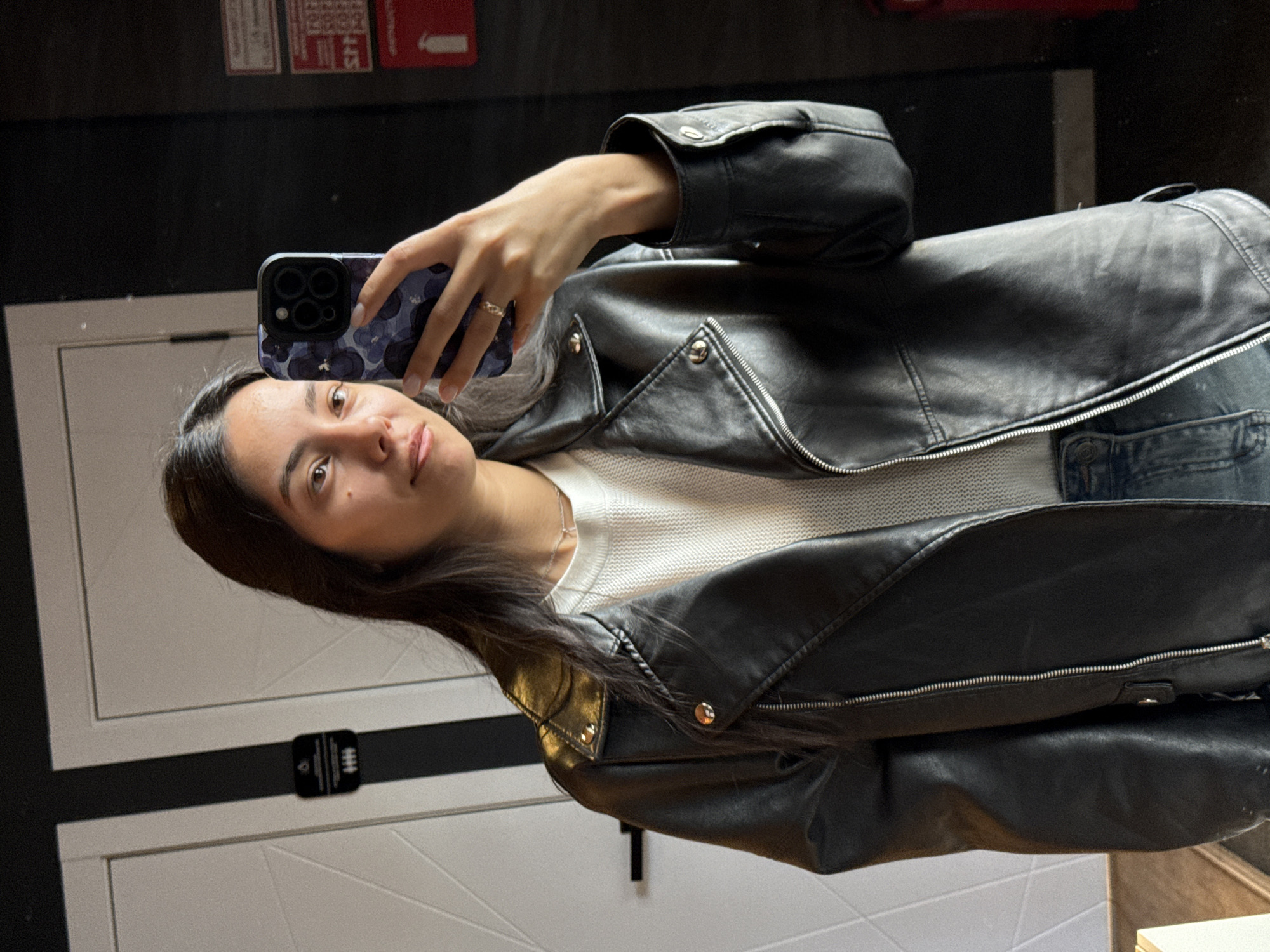 A young woman takes a selfie in a mirror. She holds a phone with a dark blue patterned case. She is wearing a white sweater and a black leather jacket. She has long dark hair. The background is a mix of dark and white walls, with a fire extinguisher sign visible at the top. A door with a black and white sign is partially visible. There is no information to verify the location in the city of Chaikovsky, Russia.