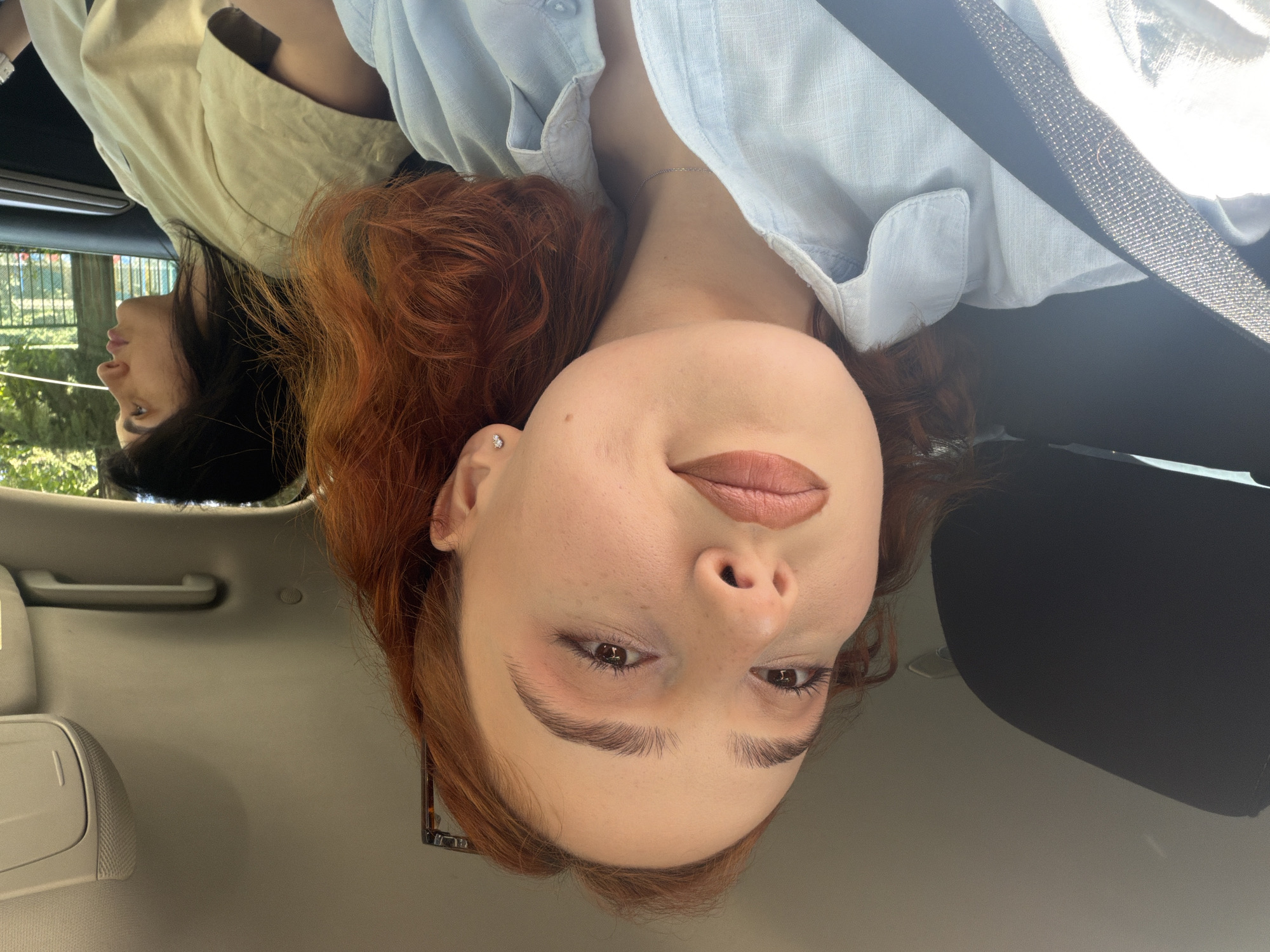 The image is taken from inside a car, showing two women. The woman in the foreground has fair skin, reddish-brown curly hair, and is wearing a light blue button-up shirt. She is positioned with her head facing the camera and tilted slightly. A black seatbelt strap crosses her chest. To the left, in the background, is another woman with dark hair, wearing a tan-colored shirt. She is looking towards the left side of the frame. The interior of the car is visible, including the headliner, a handle, and part of the dashboard. Through the car window, foliage and greenery suggest an outdoor setting. The location is likely not directly discernible from the image without further context. The location in the city of Tbilisi, Georgia is indeterminate.