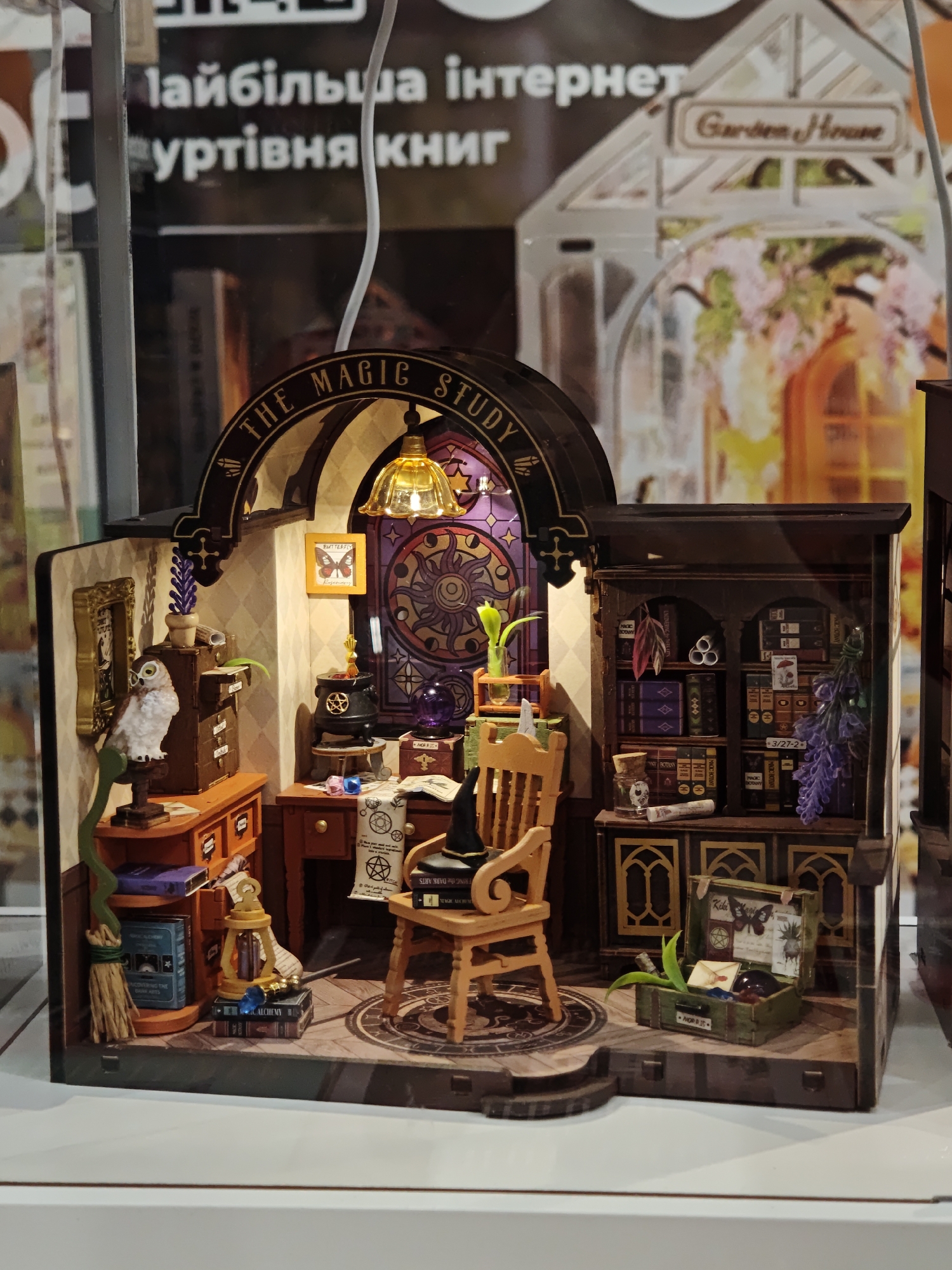 The image displays a miniature diorama of a magical study, likely a model kit or display piece, housed within a shop in Lviv, Ukraine. The diorama features an owl perched on a shelf, a desk with a lamp and cauldron, a wooden chair, and a bookshelf filled with tiny books. The background suggests a fantastical interior with stained-glass windows. The overall scene is detailed and intricate, evoking a sense of enchantment and scholarly pursuit.