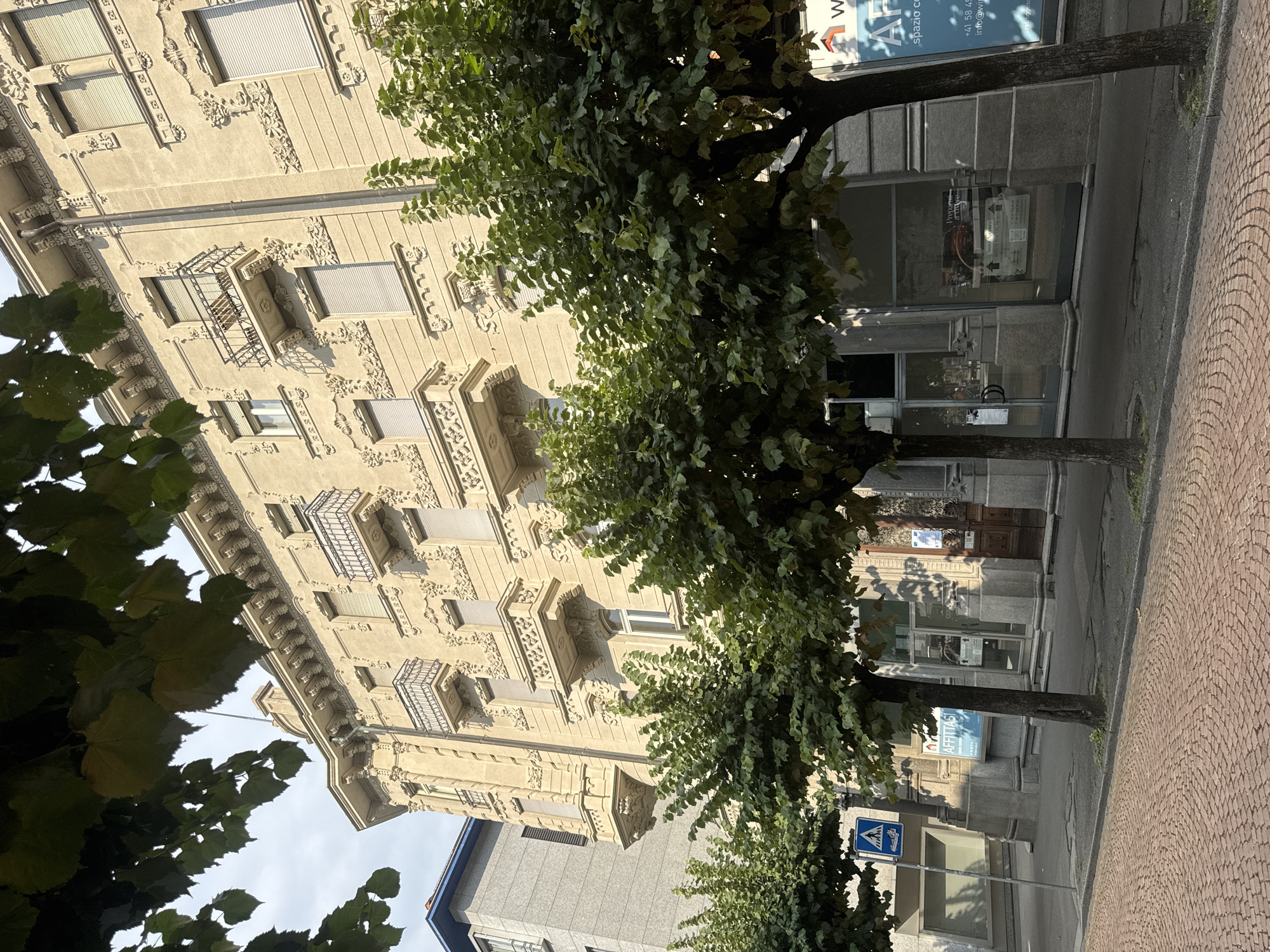 The image depicts a street scene in Bellinzona, Switzerland, featuring a multi-story building with ornate architectural details. The building, constructed with light-colored stone, exhibits numerous decorative elements on its facade, including intricate carvings and balconies with wrought-iron railings. Several trees with lush green foliage line the street in front of the building, partially obscuring the lower floors. The street is paved with reddish-brown cobblestones. A pedestrian crossing sign is visible on the left side of the image. The shops on the ground floor of the building have large glass windows, some displaying advertisements. The sky is overcast, suggesting a moderately lit day. There are no people or vehicles visible in the immediate foreground.