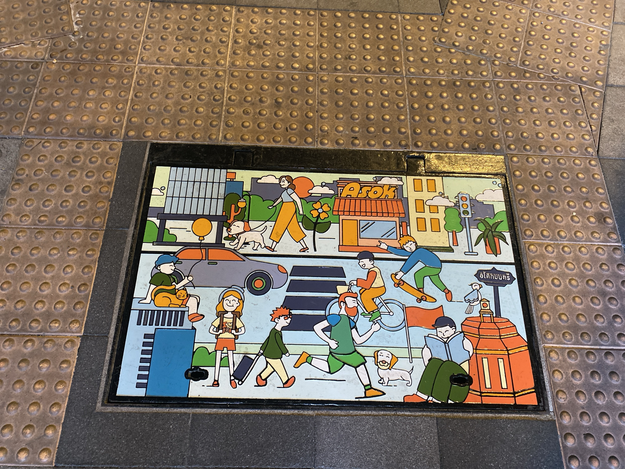 The image is a top-down view of a decorative sewer grate in the city of Bangkok, Thailand. The grate is set within a section of tactile paving, commonly used to assist visually impaired pedestrians. The paving is made of small, raised circular bumps.
The grate itself is rectangular and depicts a cartoon-style cityscape scene. The illustration features people walking, running, riding bicycles and skateboards, and interacting with pets. There are buildings, vehicles, trees, and a traffic light. One building is labeled "Asok." The illustration also includes Thai writing on a sign near an orange structure.