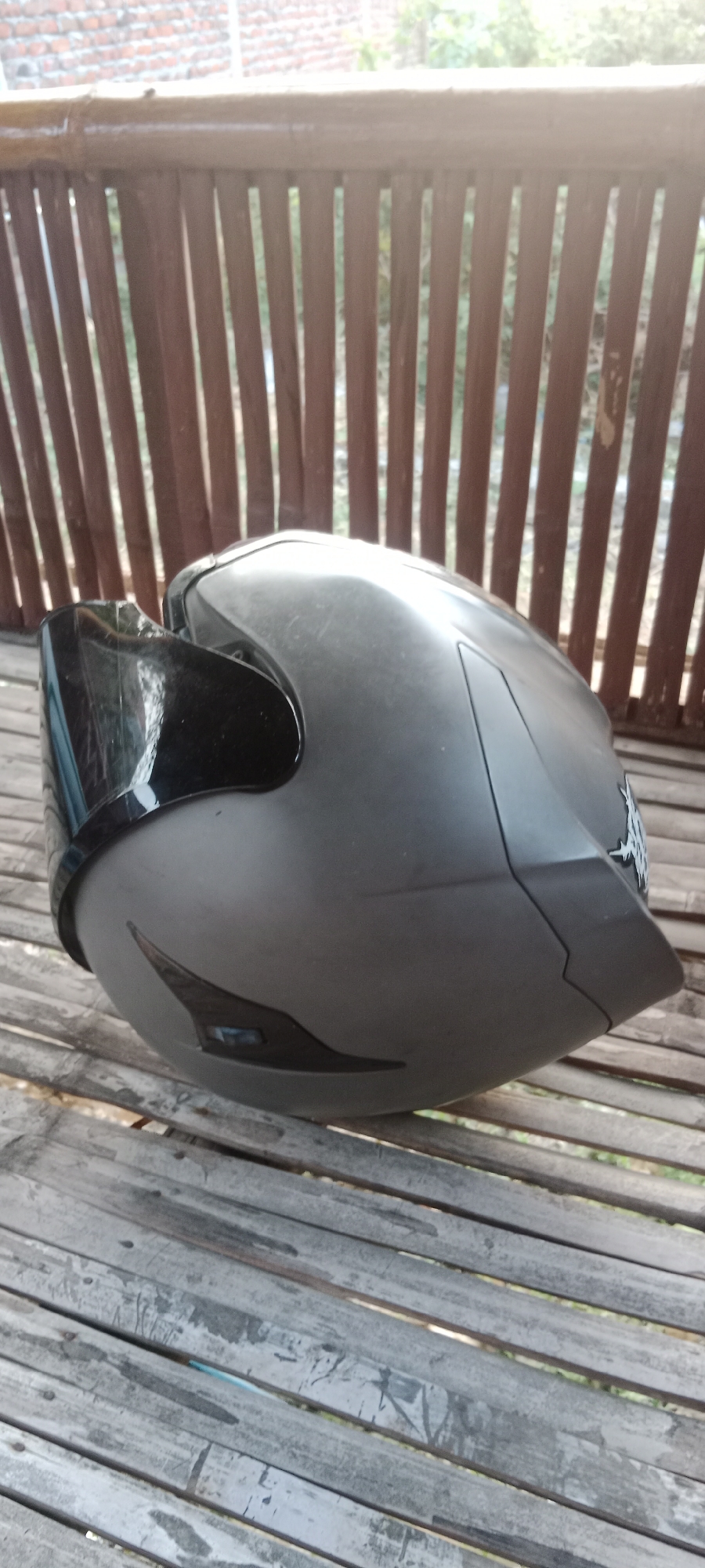 An overhead angled shot shows a matte gray motorcycle helmet resting on a weathered wooden slatted surface, likely a bench or deck, in what appears to be an outdoor setting. The helmet is positioned on its side, with the visor down. Behind the helmet, a rustic wooden slatted fence is visible, with greenery and a brick wall in the distance, suggesting a residential area in Pelang, Indonesia. The lighting suggests it is daytime, possibly late afternoon due to the strong shadows. The image captures the helmet and its immediate surroundings with a focus on textures and the play of light and shadow.
