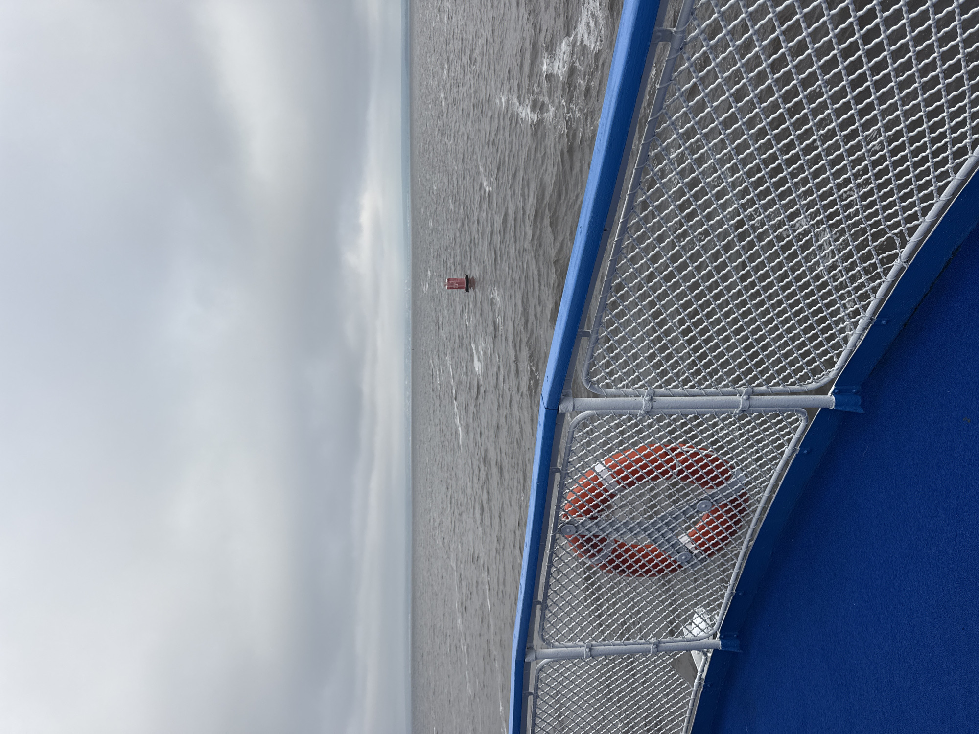 The image shows a view from the deck of a ferry, looking out over the water towards a distant, hazy shoreline. The foreground features a blue deck and a white metal railing with a blue top rail. An orange life preserver is attached to the railing. The water is choppy and gray under a cloudy sky. A red and white buoy is visible in the middle distance. The scene appears to be on the Volga River, near Laishevo, Russia. There are no people visible in the image.