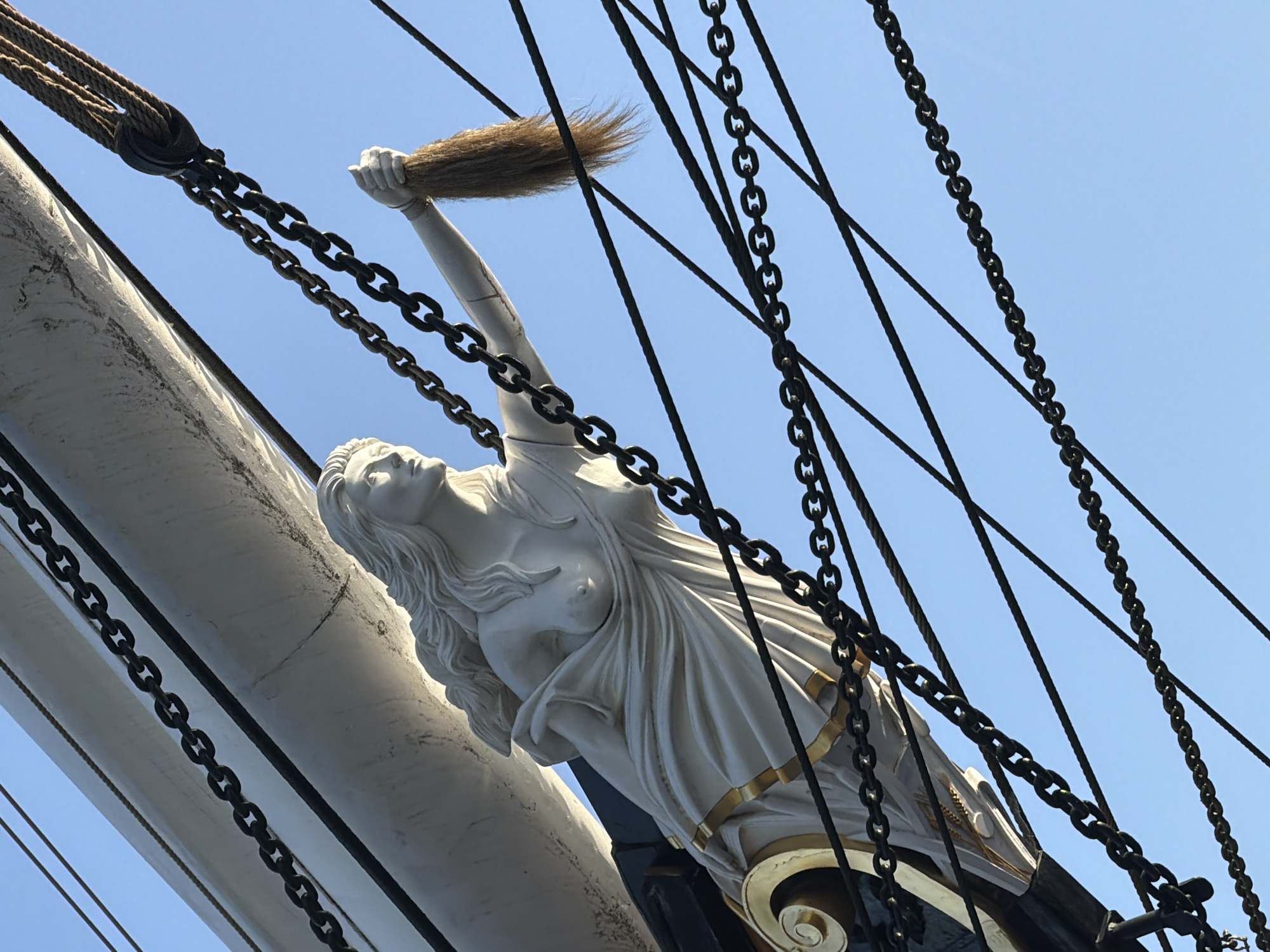 The image shows the figurehead of a ship, likely a clipper, viewed from a low angle against a clear blue sky. The figurehead is a white, classical-style sculpture of a woman, draped in fabric, with her arm raised holding a bundle of what appears to be flax or hay. The ship's hull, painted white, is visible, as are the black chains and rigging that stretch across the frame. Gold accents are visible on the base of the figurehead and the ship's structure. The photograph was taken in London, United Kingdom on the Cutty Sark.