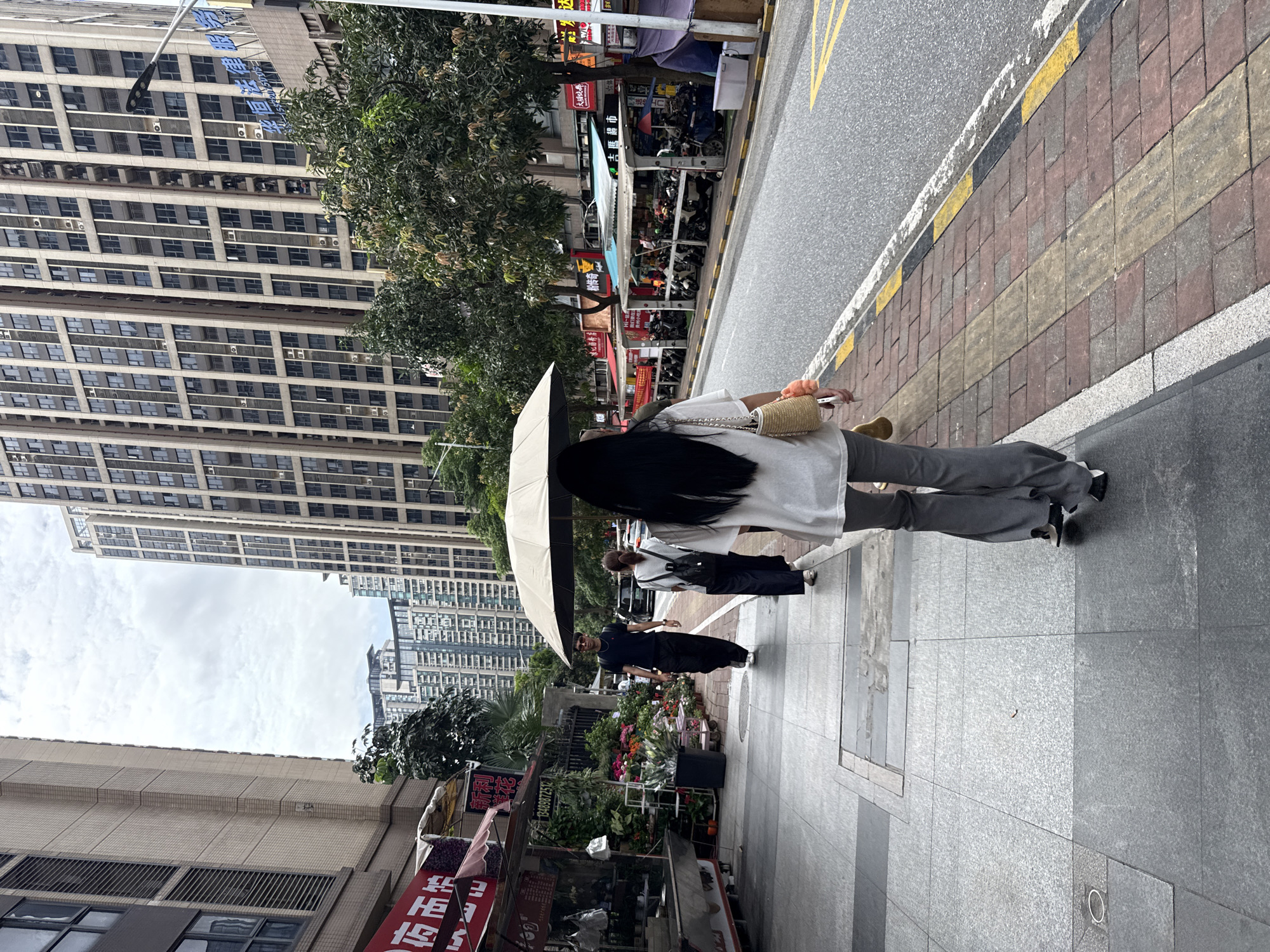 In Haizhu District, China, a pedestrian walks on a sidewalk adjacent to a street. The sidewalk is paved with gray and pink tiles, and there is a tactile paving strip for the visually impaired. A woman in a white shirt and gray pants walks away from the camera, carrying a straw bag with a fluffy pom-pom. She is holding an open beige umbrella over her head. To her left, a man in a black t-shirt and black pants walks in the same direction, wearing sunglasses. Another person, dressed in dark clothing, walks in front of the man, carrying a backpack. In the background, there are tall apartment buildings, some with balconies and air conditioning units visible. Along the street, there are several small shops and stalls, with motorcycles parked in front of them. Trees with green foliage line the street and sidewalk. The sky is cloudy.