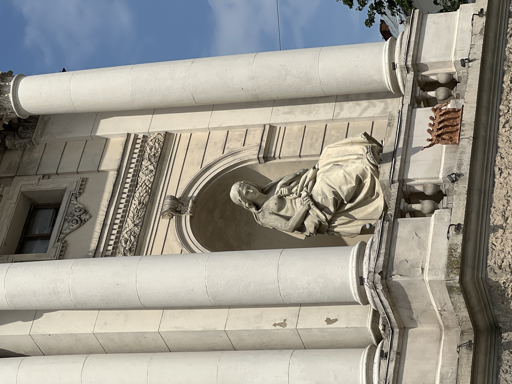 The image captures a stone sculpture of a seated female figure, likely a personification of a virtue or allegorical character, set within an alcove on the facade of a building. The sculpture is made of light-colored stone and depicts the figure draped in flowing robes, with her head bowed. She appears to be holding something in her lap. The architectural setting features large, classical columns that flank the alcove, and decorative friezes adorn the building's exterior. Below the main sculpture, a smaller group of figures, rendered in a different material and color, are visible on a ledge. The scene is lit by natural sunlight, casting subtle shadows. In the background, a clear blue sky with some clouds and the upper parts of nearby buildings are visible. The building's facade shows signs of age, with some peeling paint and weathered stone. The image appears to be taken from a slightly upward angle, looking towards the building. This is the facade of the Lviv Opera House, located in Lviv, Ukraine.