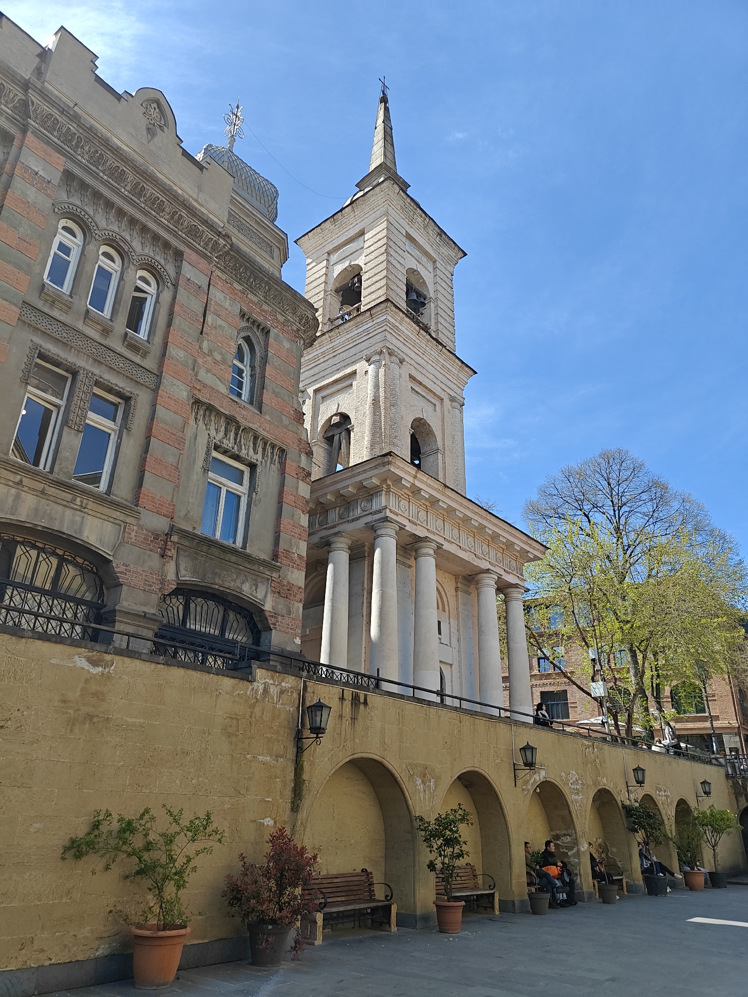 This image captures a vibrant street scene in Tbilisi, Georgia, under a clear blue sky on a bright, sunny day, likely in spring given the fresh green foliage on the trees. The composition features two distinct architectural styles and a unique tiered streetscape.
On the left, a multi-story building exhibits an older, elaborate design with alternating horizontal bands of reddish brick-like material and grey plaster. Its facade is adorned with decorative cornices, arched windows with white frames, and ornate iron grates below the ground-level windows. A small dome topped with a decorative, cross-like finial is visible on its roofline.
To the right, a prominent, light-colored bell tower rises, crowned with a spire and a cross. Bells are visible within its arched openings. Below the bell tower, a classical portico with large, smooth columns supports an entablature, indicating the entrance to a church or religious building.
In the foreground, a robust, light-brown stone wall forms a lower level, featuring a series of arched recesses. Inside these arches, several people are seated on wooden benches, relaxing or socializing. Potted plants, including small trees, are placed both within and outside these arches. Black, ornate street lamps are affixed to the wall above the arches. Above this lower wall, an ornate black metal railing separates the street level from a raised platform where the church entrance is located. A large tree with bright green, new leaves stands beside the church, adding a touch of natural beauty to the urban setting. Another person can be seen on the raised platform near the church. No discernible text is visible in the image.