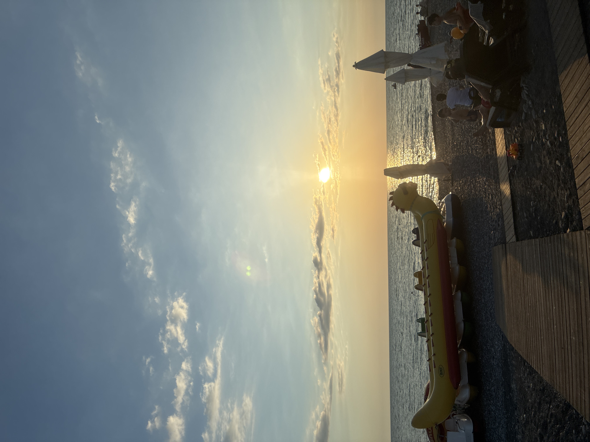 This image depicts a sunset on a beach in Sochi, Russia. The sun is low in the sky, casting a warm, golden glow over the scene and reflecting on the water. In the foreground, a wooden walkway leads to the pebbled beach. A yellow inflatable banana boat, designed to resemble a dragon, is prominently displayed on the beach, along with other inflatable watercraft. Several beach umbrellas are visible, some in their closed state. A few people are scattered across the beach, with some seated on lounge chairs and others standing. The sea is calm, with gentle waves rolling towards the shore. The sky is a gradient of blue, transitioning to orange and yellow near the horizon, with scattered clouds catching the light.