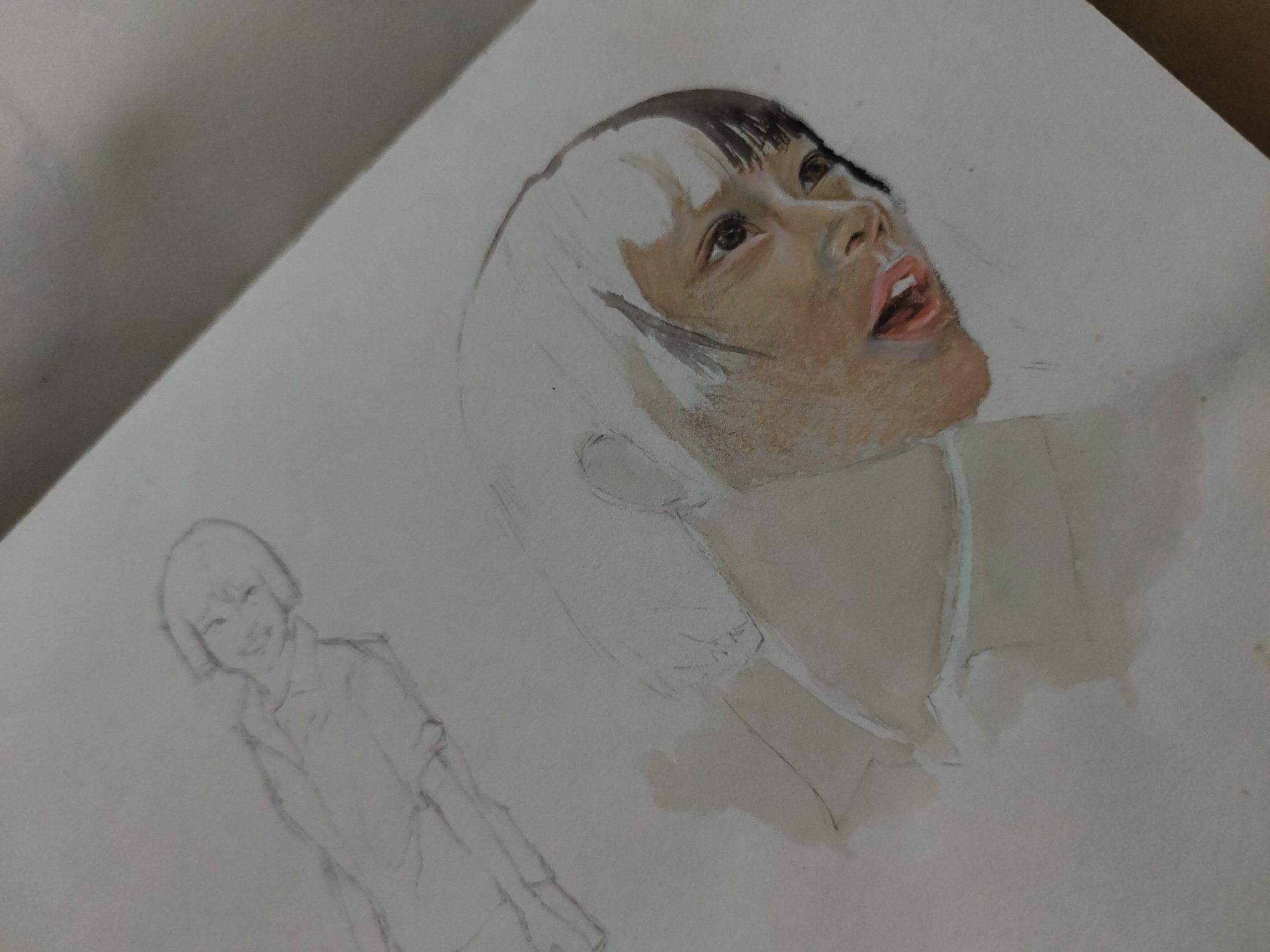 The image displays an in-progress artwork on a light-colored paper sheet, likely from a sketchbook, viewed from an angled, slightly elevated perspective. The primary subject is a partially colored portrait occupying the upper right portion of the page. This portrait features a person, likely female, with dark, straight bangs and a prominent, unpainted or light-colored section of hair on the left side of their head. The subject's face is rendered with warm, medium-brown skin tones, particularly around the cheeks and jawline, contrasting with uncolored areas on the forehead and the main body of the hair, which are delineated by pencil lines. The eyes are dark and gaze upwards. The mouth is open, revealing a pink lower lip and tongue, suggesting an expression of awe, singing, or surprise. The neck and shoulders are outlined in pencil, with a faint wash of color indicating a collar or clothing. To the lower-left of the main portrait, a smaller, uncolored pencil sketch of a smiling young person with short hair and a collared shirt is visible. The background is the plain paper, showing minor smudges and faint pencil marks. No elements within the image indicate a specific geographical location such as Davao City, Philippines.