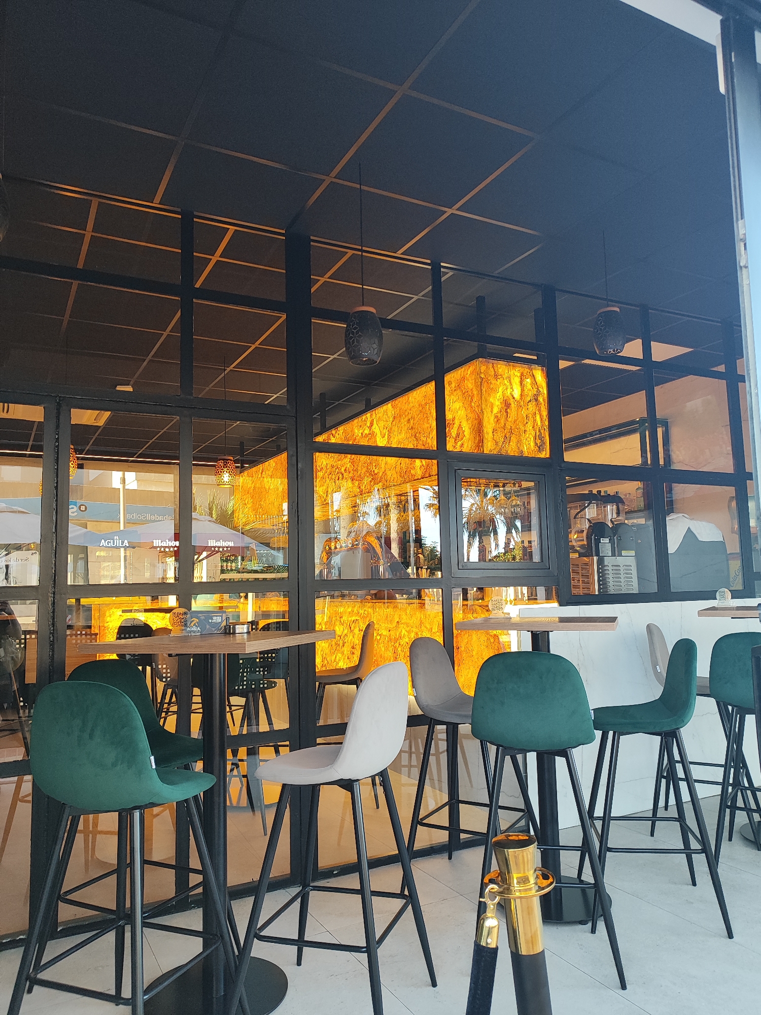 The image depicts the interior of a modern establishment, likely a bar or restaurant, with large glass windows that offer a view of the street and reflect the warm, golden light emanating from within. The interior features high tables and bar stools in green and gray upholstery, arranged in front of a distinctive wall adorned with backlit, textured panels that resemble polished stone or amber, casting a vibrant, glowing ambiance. The ceiling is a dark grid structure, and decorative pendant lights hang from it. In the foreground, a golden stanchion with a black velvet rope stands on the tiled floor. The reflections in the glass show outdoor umbrellas with "AGUILA" and "Mahou" branding, suggesting a location in Spain. Specifically, the establishment is located on Calle del L'Alcalde, El Campello, Spain, as indicated by the visible signage. There are no identifiable people in the image.