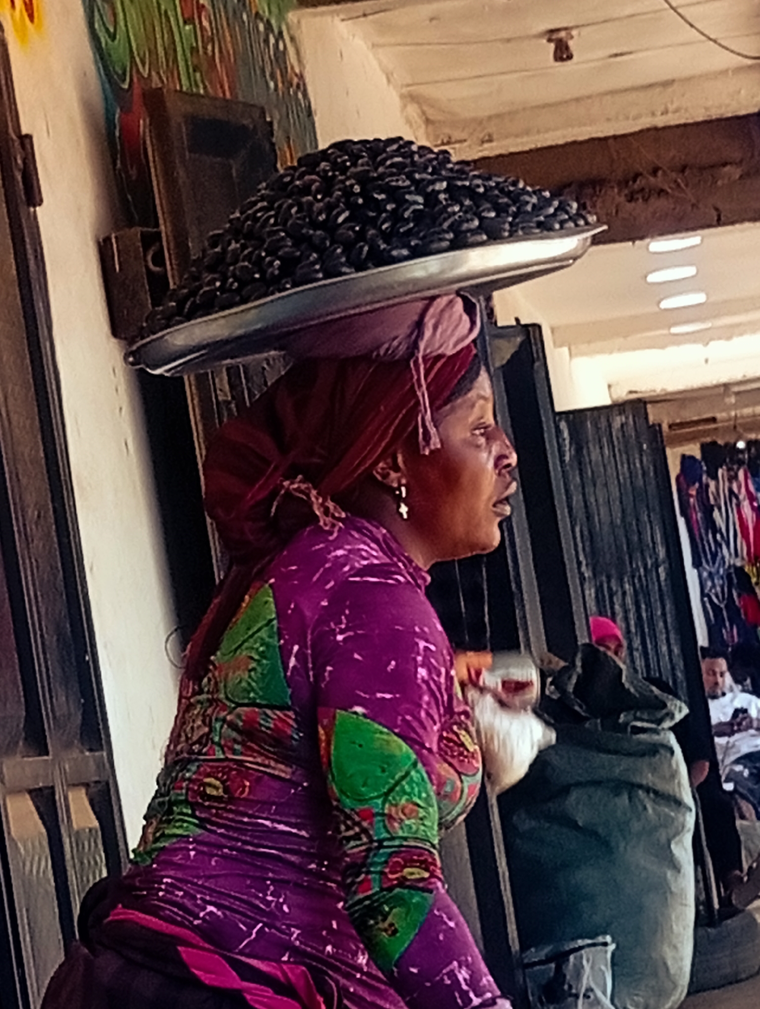 A woman is photographed in Bukuru, Nigeria, carrying a large platter of black olives on her head. She is wearing a vibrant purple and green patterned dress and a maroon headscarf. Her mouth is slightly open as if speaking or reacting to something. Behind her, a large, dark green sack is visible, and further in the background, a man in a white t-shirt stands near a market stall displaying colorful garments. The environment appears to be an outdoor marketplace, with a rough, textured wall and a corrugated metal roof overhead.