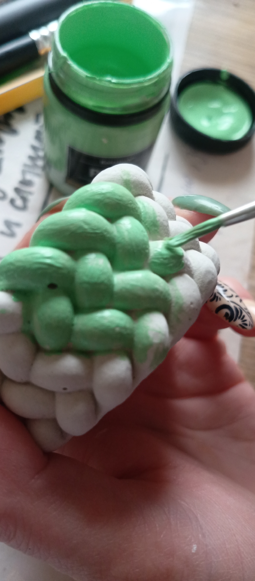 A human hand holds a textured object resembling a woven basket, being painted with green paint using a paintbrush. A jar of green paint with the lid next to it is in the background. The location is unknown.