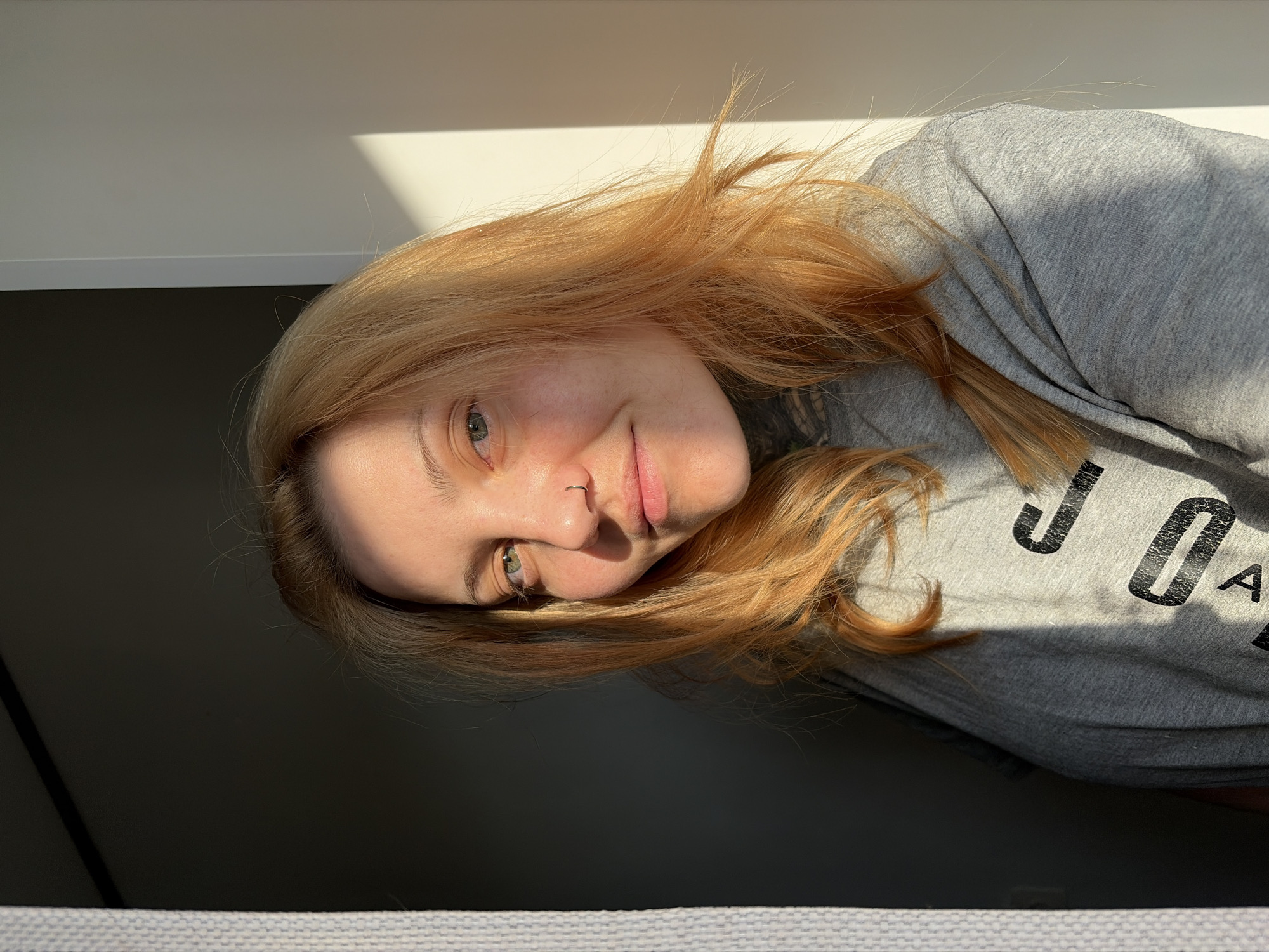 A young woman with fair skin, light green eyes, and reddish-blonde hair poses for a close-up shot. She is wearing a grey sweatshirt with black lettering. A silver nose ring is visible in her left nostril. The woman is illuminated by sunlight, creating strong shadows on her face and clothing. The background consists of a white wall with a shadow and a dark grey area. The bottom of the image shows a textured grey surface. Based on the sweatshirt and the general style, it appears that the photo was taken indoors. There is no contextual information in the image to confirm the location as Chaikovsky, Russia.