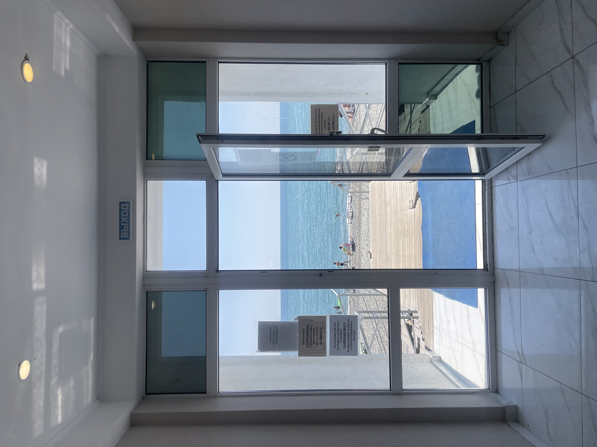 The image shows a wide glass door and window system, opening onto a view of the Black Sea. The door is slightly ajar, revealing a wooden deck and a pebble beach where a few people are visible. The water is a calm blue, with small waves. The sky is clear and bright. Above the door, a blue sign reads "ВЫХОД" (EXIT) in white letters. Inside, the floor is tiled with large white tiles with grey veins. There are also several notices posted on the glass panes, two of which are legible. One notice states that the hotel's beach is open from 06:00 to 00:00. Another notice, in Russian, informs guests that smoking is prohibited on balconies and in rooms, and that they may only smoke in designated areas. The location is identified as Giachrypsh, Abkhazia.