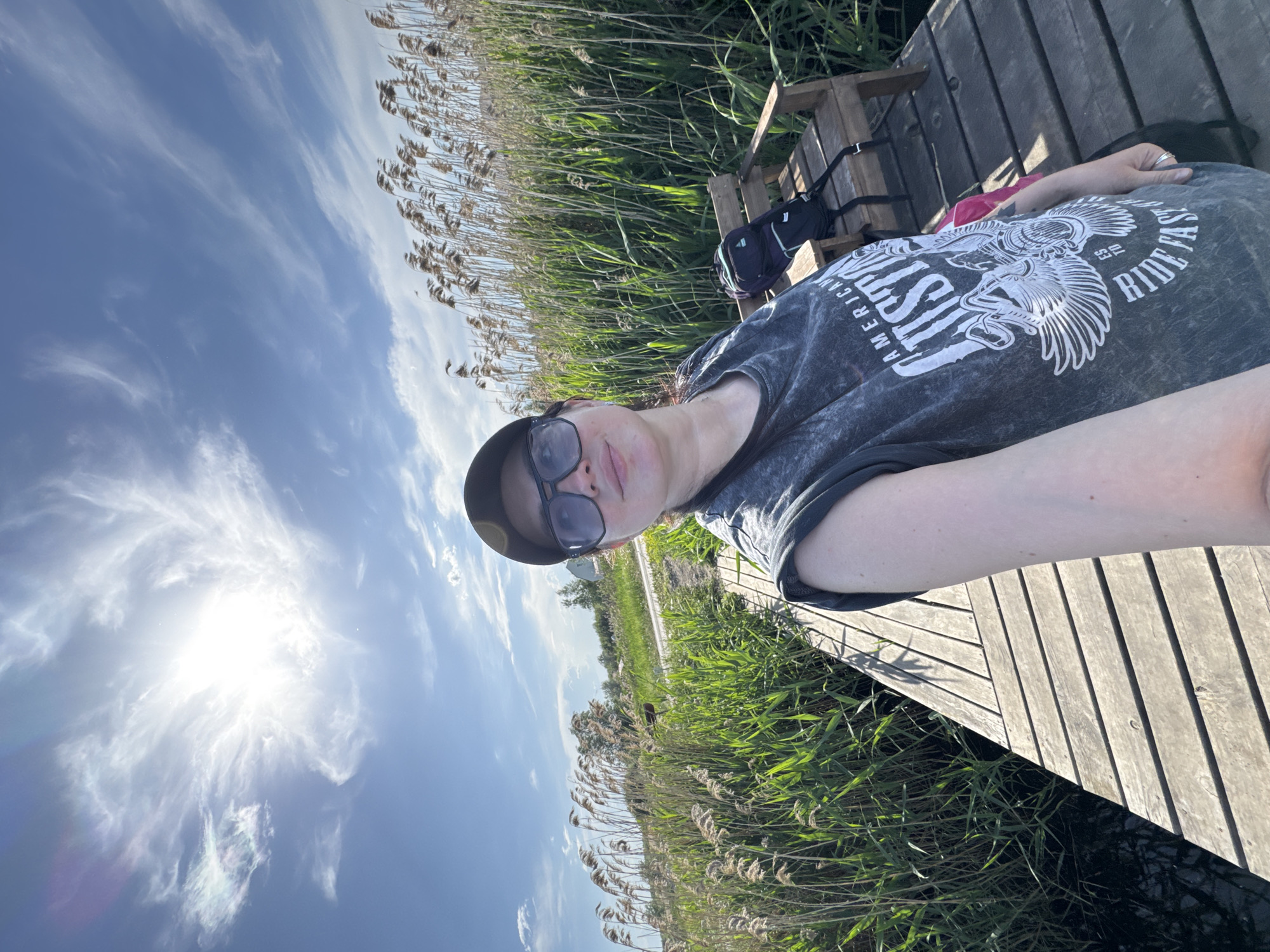 A woman lies on a wooden platform next to tall grasses or reeds. She wears sunglasses and a cap. A backpack sits on a wooden bench behind her. The sky is bright with scattered clouds and the sun shining. The location appears to be outdoors, possibly near a body of water with the reeds growing along the edge. The city and specific details about the location are not discernible.