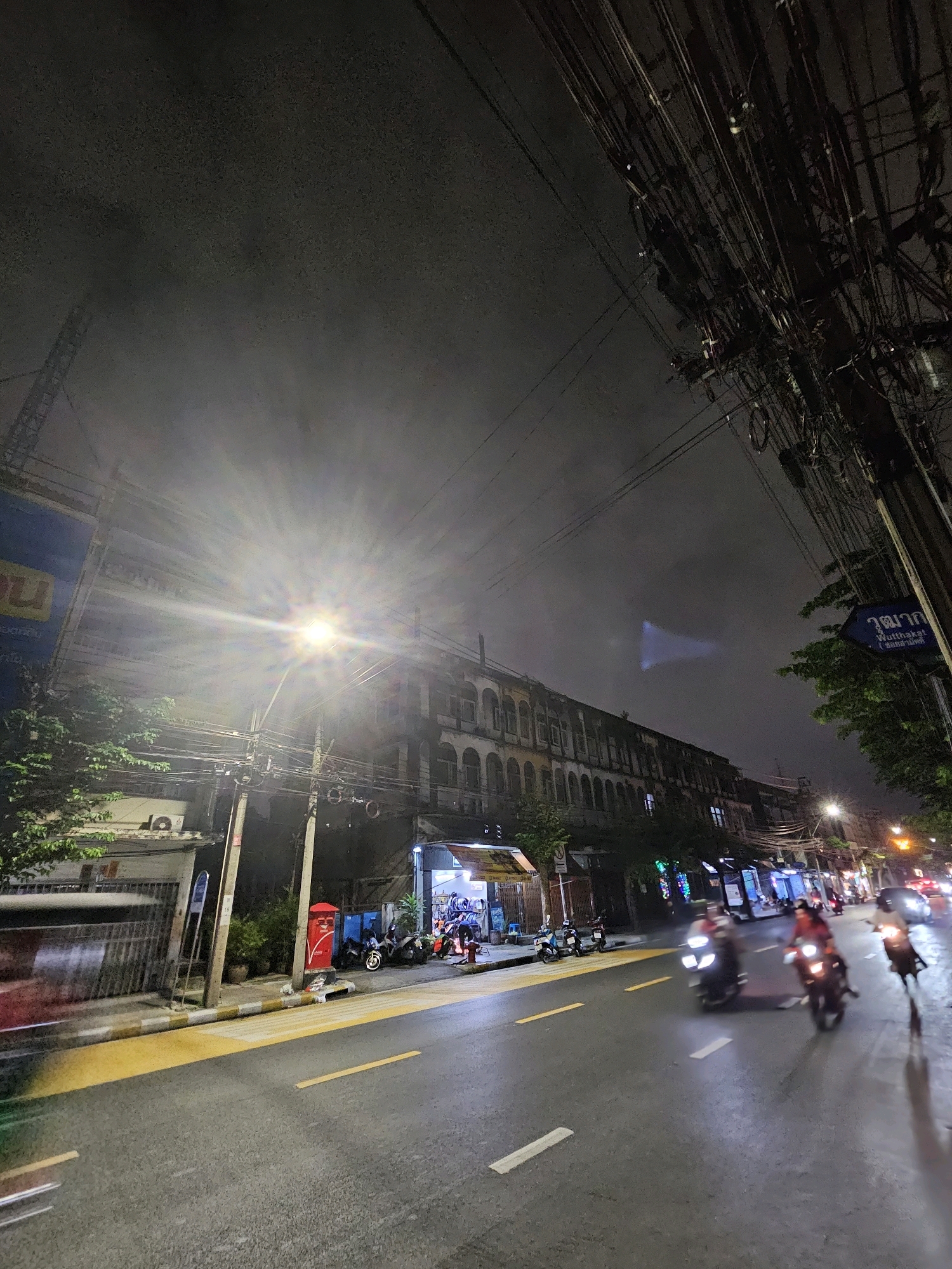 This is a photograph taken at night on a city street in Bangkok, Thailand. The street is wet, suggesting recent rain, and illuminated by streetlights and the headlights of passing vehicles. On the right side of the street, there are buildings with shops or residences, and a dense network of electrical wires stretches overhead. Several motorcycles are visible on the street, some parked and others in motion, with their headlights on. A few people are seen riding the motorcycles. The sky is dark and hazy. The overall impression is of a busy urban environment at night.