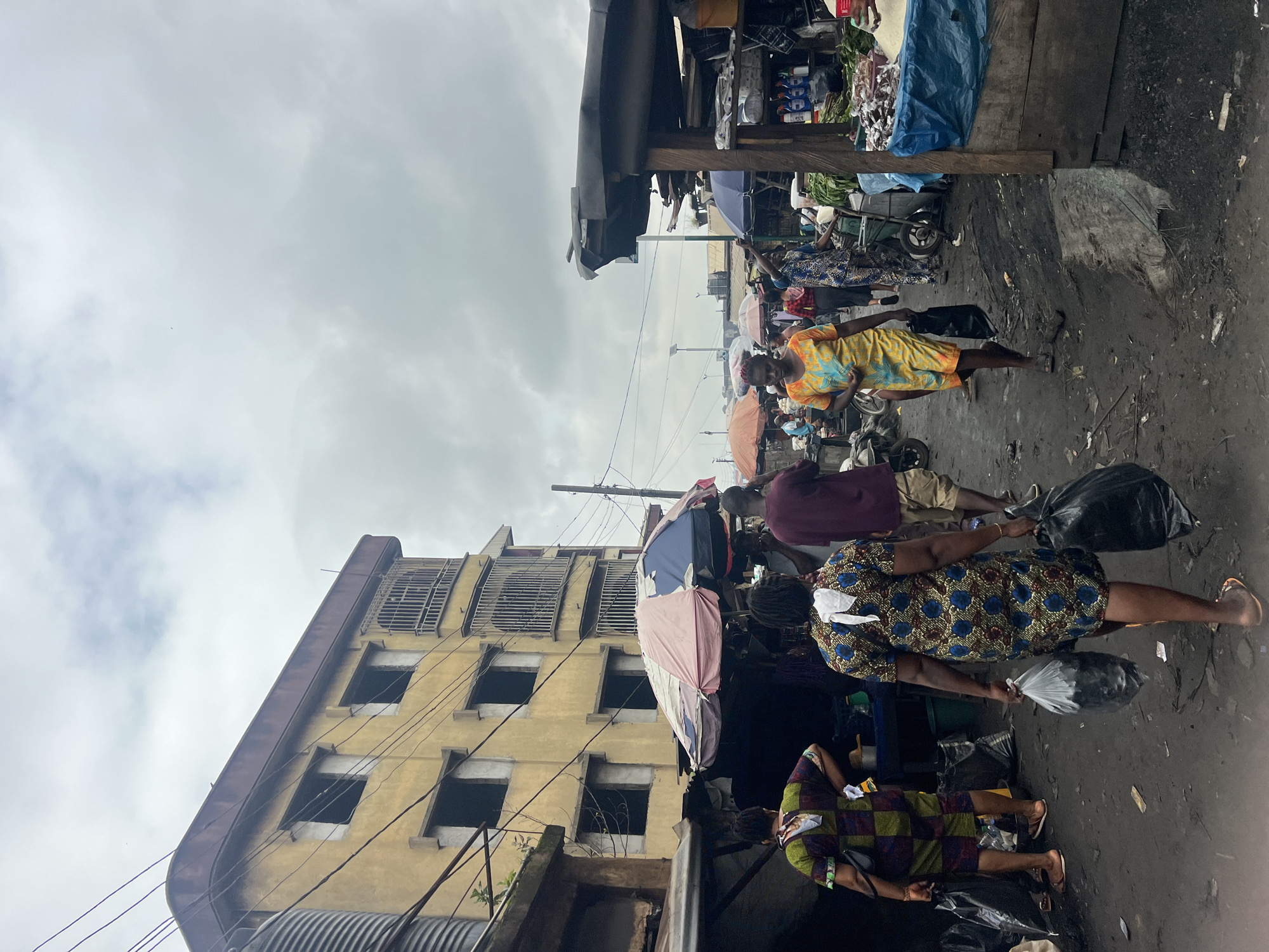 The image depicts a busy street market scene in Nkpor, Nigeria. In the foreground, several individuals are walking through the dusty market lane. One woman in a blue and gold patterned dress carries two black plastic bags, one in each hand, as she walks away from the camera. To her left, another woman in a colorful checkered outfit walks away from the camera, also carrying a black plastic bag. Further in the background, more people are visible, some interacting with market stalls.
The market is lined with temporary structures, many covered with tarps and umbrellas, displaying various goods. A prominent feature of the scene is a multi-story, weathered yellow building on the left side of the image, appearing partially dilapidated with some windows boarded up. Overhead, a network of electrical wires crisscrosses the sky. The ground of the market is uneven and covered in dirt and debris. The sky is overcast, with a muted, cloudy atmosphere. The overall impression is of a vibrant but somewhat unkempt urban marketplace.