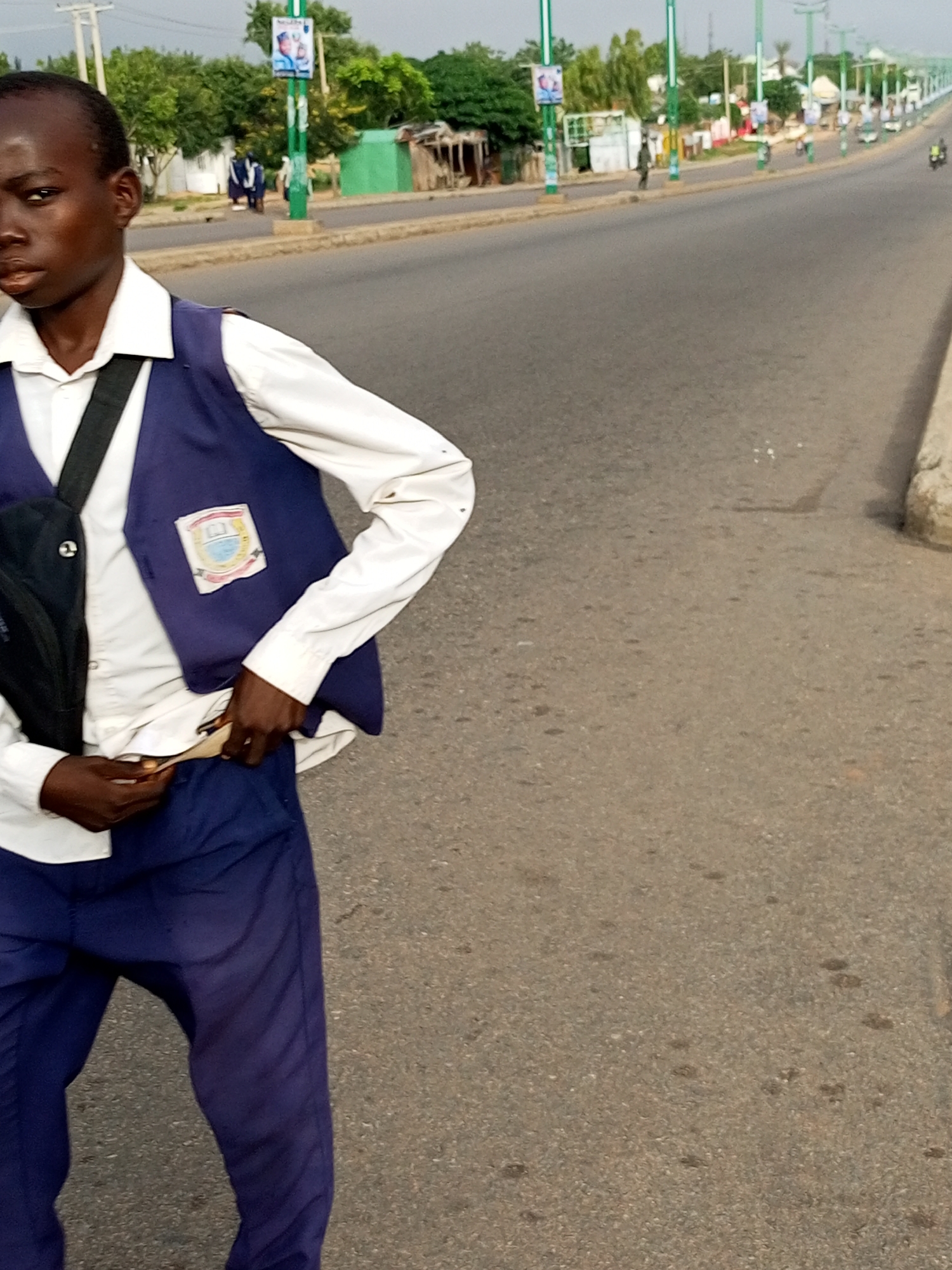 A young boy, appearing to be of school age, stands on a paved road in Gombe, Nigeria. He is wearing a white collared shirt, a dark blue vest with a school crest on the left side, and dark blue trousers. The boy has a black bag slung across his left shoulder and is holding a piece of paper or fabric tucked into his trousers with his right hand. The road is asphalt and appears to be a multi-lane thoroughfare with streetlights and promotional posters lining the side. In the background, there are trees, buildings, and other people walking and vehicles moving in the distance. The overall lighting suggests it is daytime.