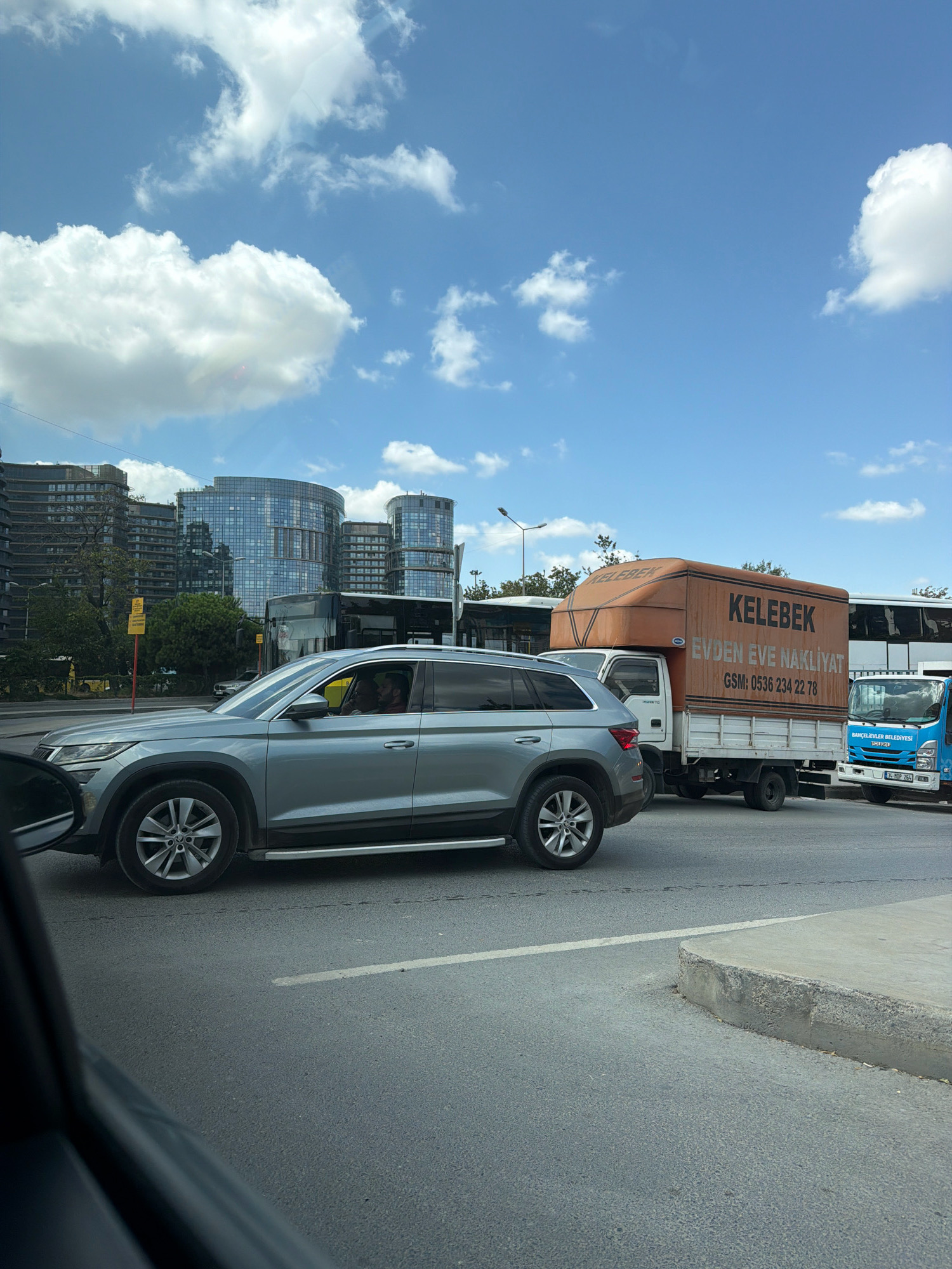 The image shows a street scene in Bakırköy, Turkey, on a sunny day with scattered clouds. In the foreground, a silver SUV is stopped at an intersection, with its driver visible. Behind the SUV, a large orange truck with the text "KELEBEK EVDEN EVE NAKLİYAT" (Butterfly Home to Home Moving) and a phone number is also stopped. To the right of the orange truck, a smaller blue Isuzu truck is visible. In the background, several modern buildings with glass facades line the street, suggesting an urban commercial or residential area. The road surface is asphalt with painted lane markings.