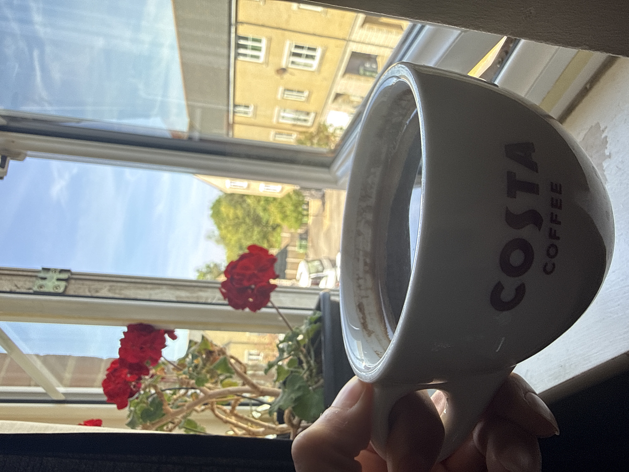 A hand holds a Costa Coffee mug nearly empty of coffee against a window. Outside the window is a view of buildings, a tree, and a red flowering plant. The mug is white with the Costa Coffee logo. The window is framed in white and is open. The scene is bathed in natural light. There is nothing in the image to suggest that the location is Great Yarmouth, United Kingdom.