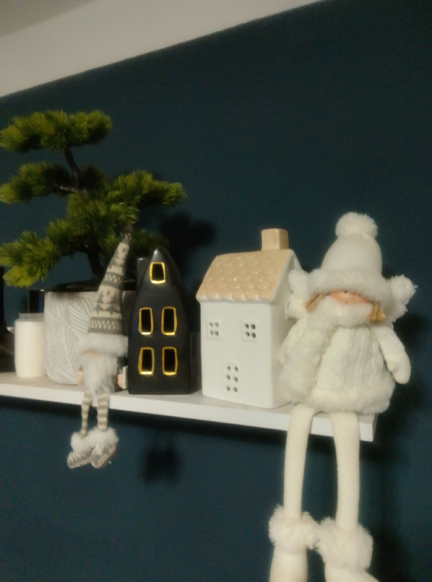 The image shows a shelf with Christmas decorations against a dark teal wall. On the left is a small artificial tree in a patterned pot. Next to it is a grey and white gnome decoration with a striped hat and fuzzy feet. To the right of the gnome is a black house-shaped decoration with gold window cutouts. Next to the black house is a white house-shaped decoration with a tan roof and small square window cutouts. Finally, on the far right, is a plush doll wearing a white winter outfit with a hat, scarf, and fuzzy trim. The doll has long legs that hang over the edge of the shelf. The overall scene is a still life display of holiday decor. There is no indication of a location in Wedel, Germany.