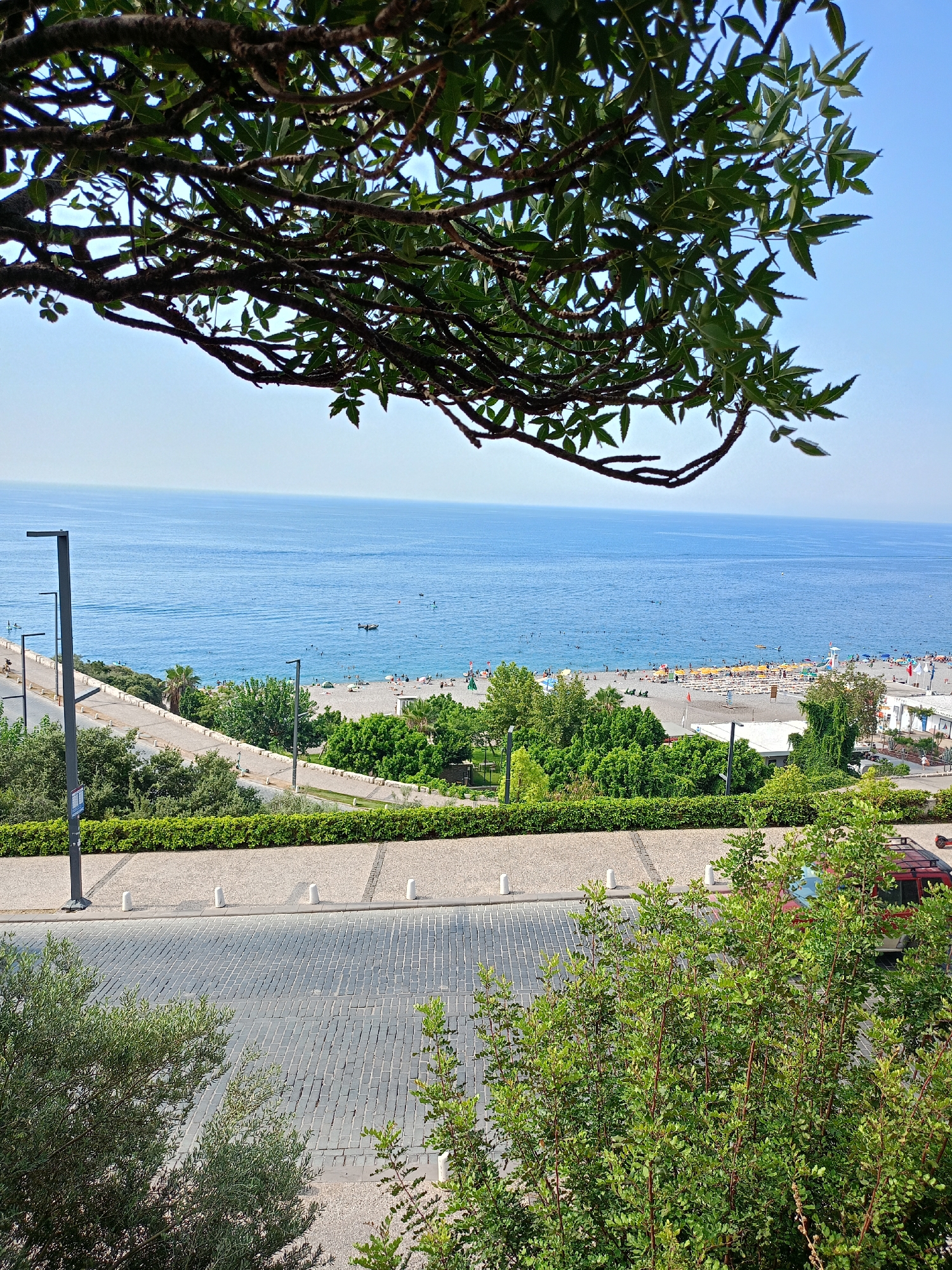 This image captures a vibrant coastal scene in Antalya, Turkey. In the foreground, a cobblestone road curves gently, bordered by lush green bushes and trees. Two modern-style lampposts stand sentinel along the road. Beyond the road and a neatly manicured hedge, a sandy beach stretches out, dotted with colorful umbrellas and sunbathers. The azure Mediterranean Sea serves as a breathtaking backdrop, with a few small boats visible on its surface and people enjoying the water. The clear blue sky overhead suggests a warm, sunny day. The overall impression is one of a lively and picturesque holiday destination.