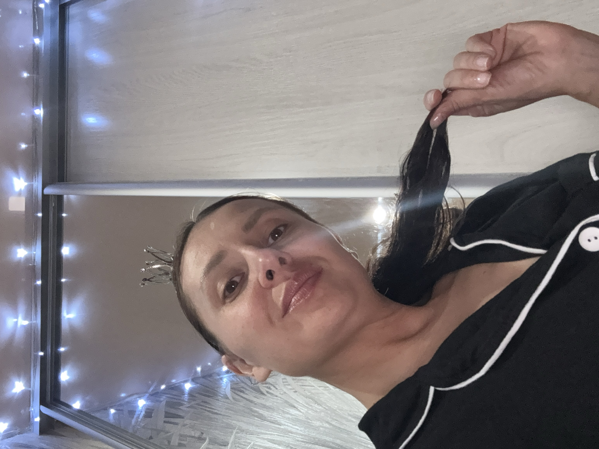 The image displays a woman's face and upper torso, captured from a low, tilted angle, suggestive of a selfie taken in a mirror. The woman has dark hair and a faint smile, looking towards the camera. A small, metallic tiara rests on the left side of her head. She is wearing a black top, possibly pajamas, detailed with white piping and a white button visible on her right shoulder. Her right hand is seen holding a section of her dark hair.
The background features bright, white or bluish fairy lights arranged vertically along a structural frame, casting soft illumination on a light-colored wooden or laminate surface. Below the woman's head, there is a substantial amount of white, fluffy or stringy material, likely part of decorative elements or bedding. The lighting implies an indoor setting, potentially at night, with the fairy lights providing the main source of light. The location context is Chaikovsky, Russia. No text is visible in the image.