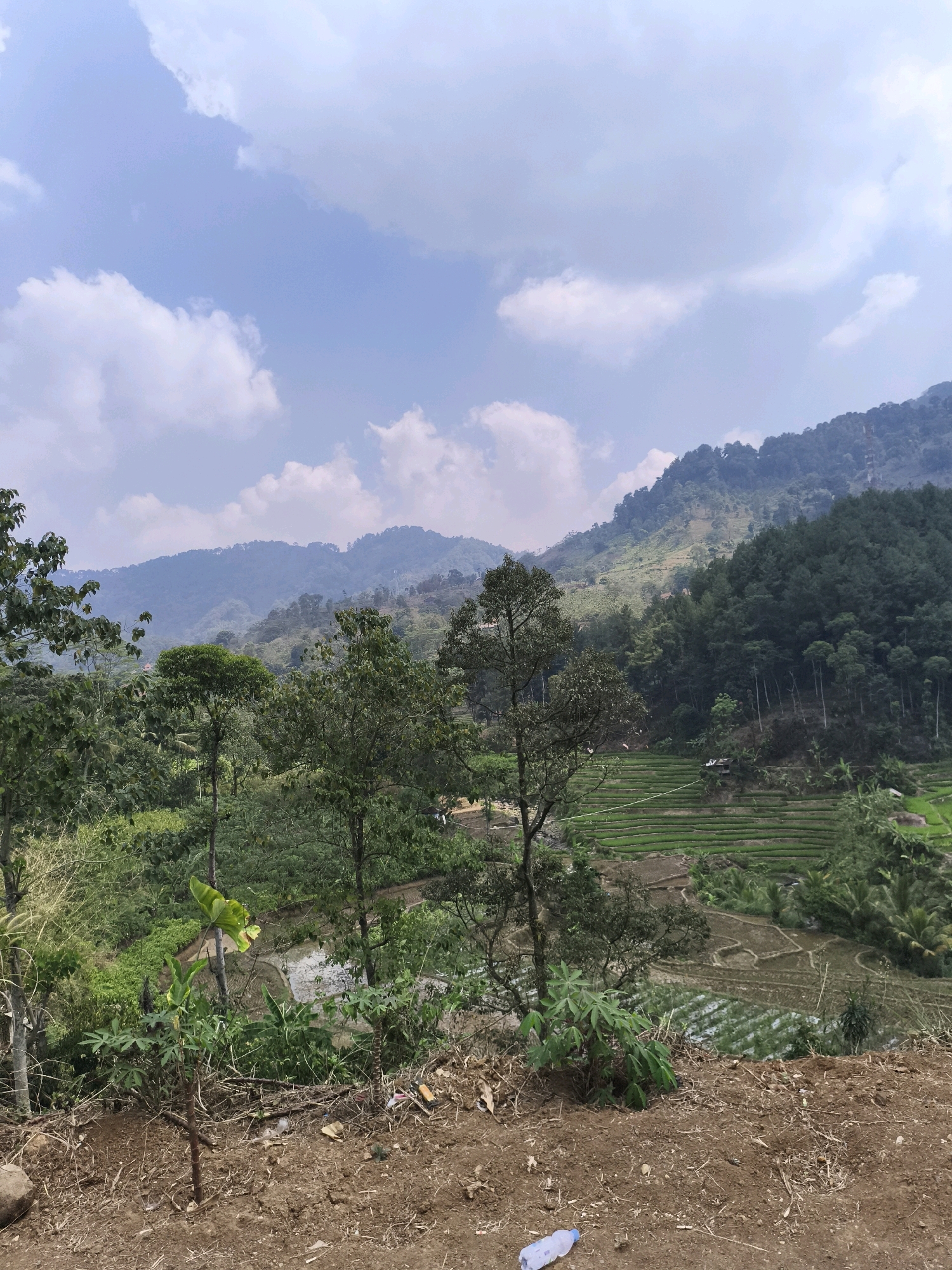 This is a landscape photograph taken in Babakan Madang, Indonesia. The scene depicts rolling hills covered in lush greenery, with a prominent vista of terraced rice paddies in the mid-ground. The sky is partly cloudy, with soft white clouds drifting across a pale blue expanse. In the foreground, dry, brown soil dominates, with some sparse vegetation and fallen twigs. A discarded plastic water bottle lies on the soil in the lower right corner. Beyond the rice paddies, dense forests of various trees cover the slopes of the mountains, with some lighter patches indicating human settlement or different vegetation types. The overall impression is one of a rural, agricultural landscape with natural beauty.