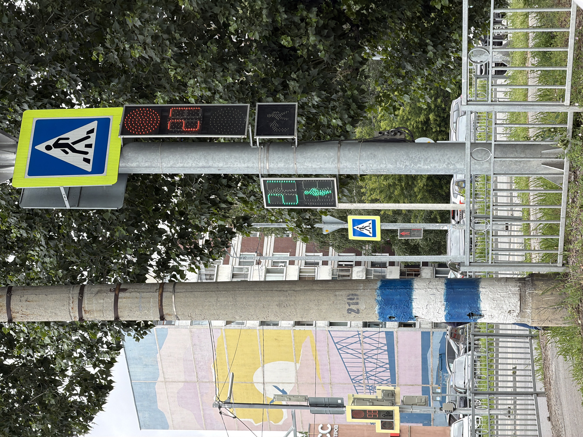 The image depicts a street scene in Ulyanovsk, Russia, featuring a concrete utility pole with blue and white stripes. Attached to the pole are traffic signals and pedestrian crossing signs. A pedestrian crossing signal displays a red walking figure with a countdown timer showing "27". Another signal shows a green walking figure. A pedestrian crossing sign with a blue background and a white silhouette of a walking person is visible. In the background, a multi-story apartment building with a colorful mural on its facade stands. The mural depicts a pink and yellow abstract landscape with a bridge motif. Other buildings and trees are also present, along with parked cars on the street. A metal fence lines the sidewalk.