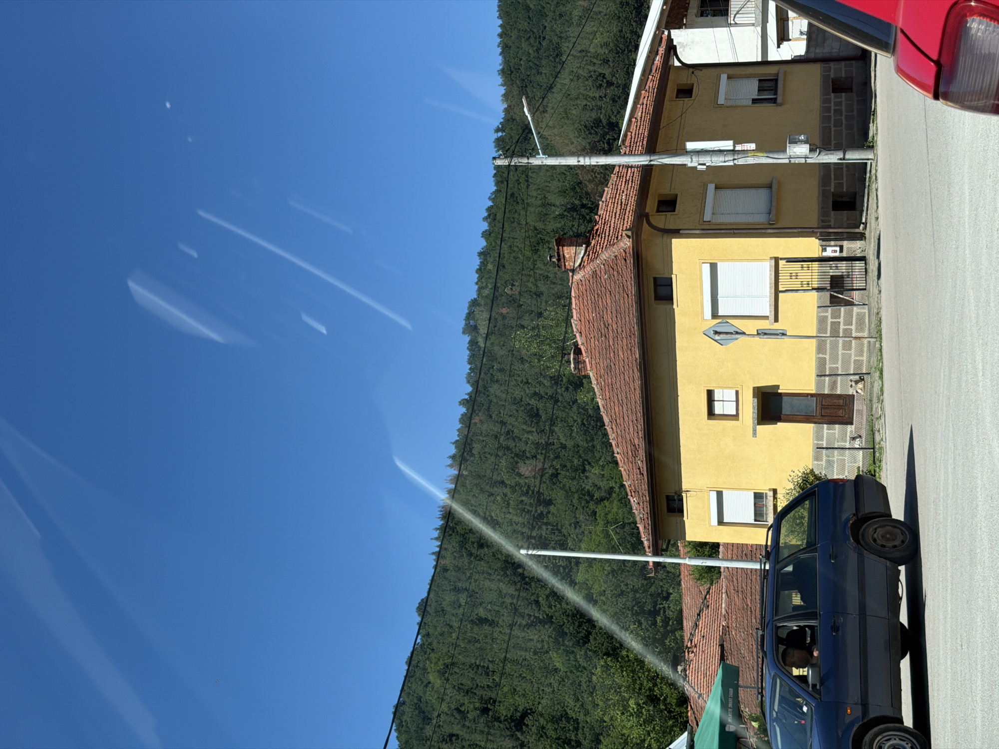 In Beli Osam, Bulgaria, under a clear blue sky, a bright yellow house with a terracotta tiled roof stands prominently. The house has several windows, some with white rolling shutters, and a wooden door. A street lamp is affixed to a utility pole near the house. In the foreground, a dark blue station wagon is parked, with a driver visible inside. A partial view of a red vehicle is also present on the right side of the image. Behind the house, a dense green forest covers a hillside, with a thin white stream of water arcing across the sky from the left, originating from a pipe extending from beyond the frame and falling towards the hillside. The scene appears to be a residential area with a natural backdrop.