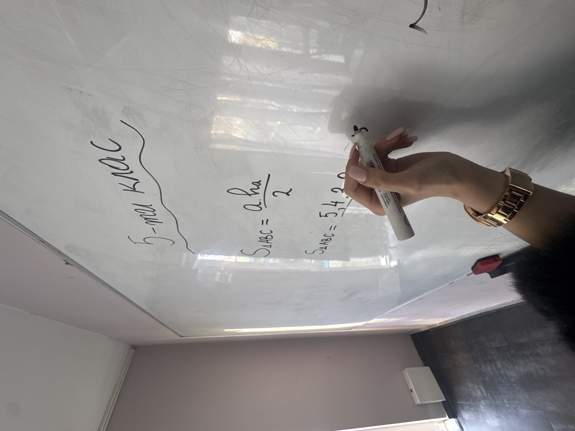 The image shows a person writing on a whiteboard in a classroom setting, likely in Sofia, Bulgaria (based on the Cyrillic alphabet used in the text). The person, wearing a gold watch, is using a marker to write mathematical formulas. The whiteboard has the inscription "5-ти клас" (meaning "5th grade"). Below this are written formulas for the area of a triangle: "S△ABC = a*ha/2" and "S△ABC = 54,2". A whiteboard eraser is visible on the right edge of the board. The classroom walls are painted in light colors.